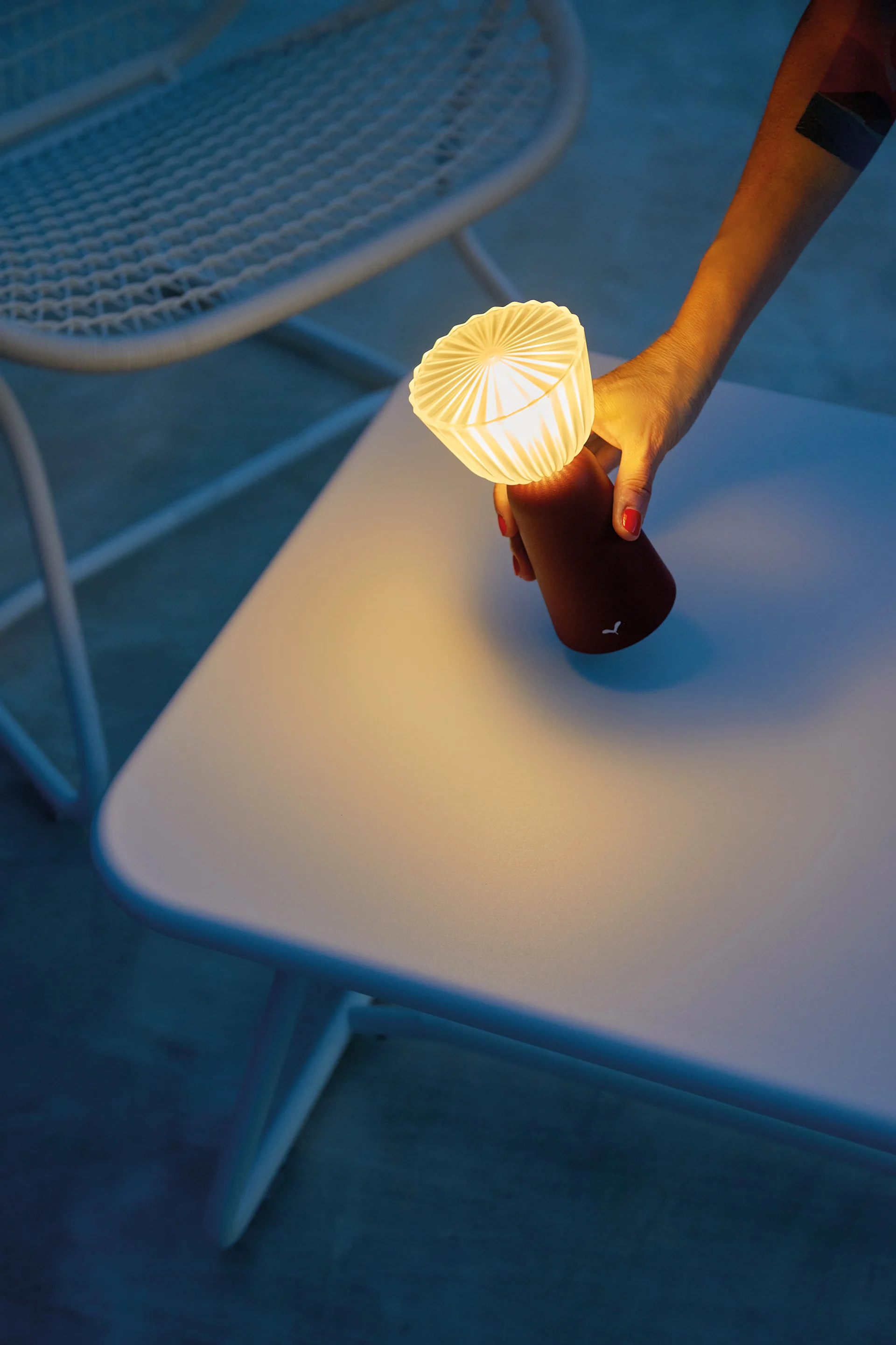 Hand mit rotem Nagellack hält eine kleine leuchtende tragbare Lampe mit plissiertem Oberteil über einem hellen Tisch.