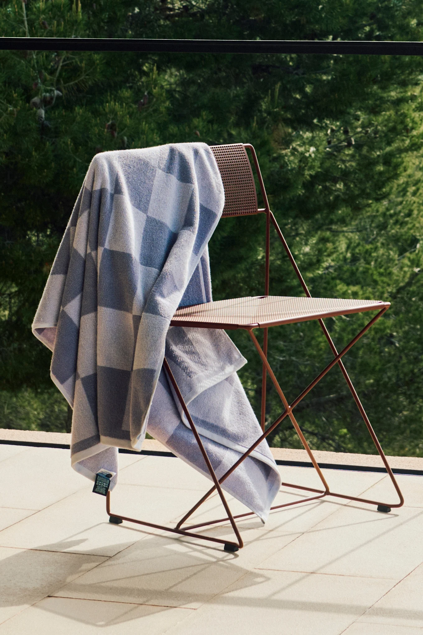Hellblaues und graues kariertes Handtuch auf einem rotbraunen Metall-Klappstuhl auf einer sonnigen Terrasse mit grünem Laub.