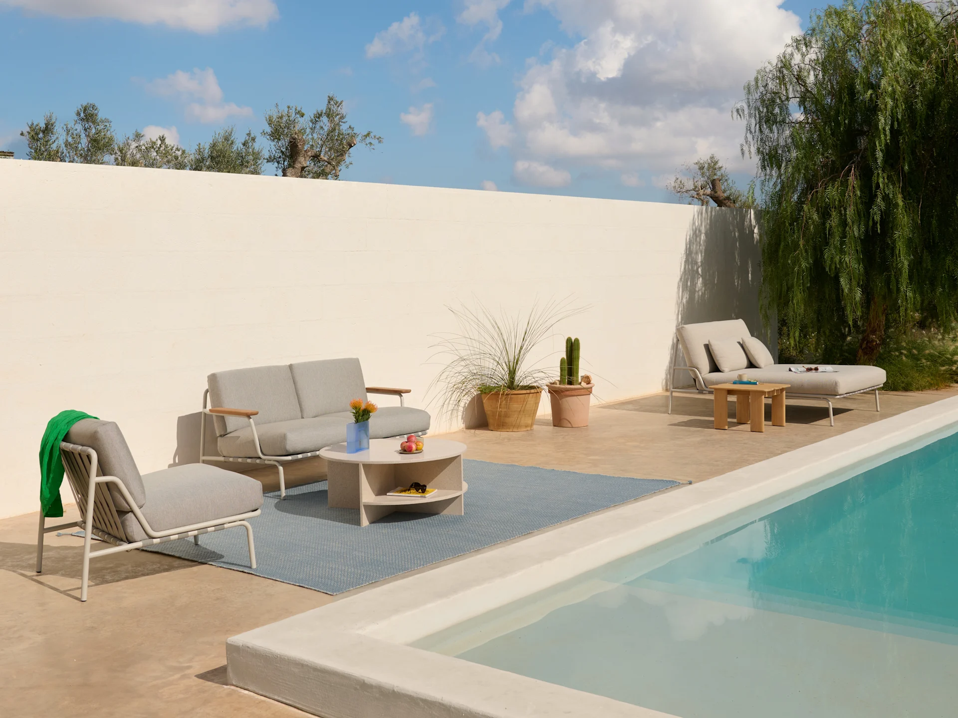 Außenterrasse mit hellgrauem Sofa, Sesseln, Couchtisch auf blauem Teppich und einer Liege am Pool.