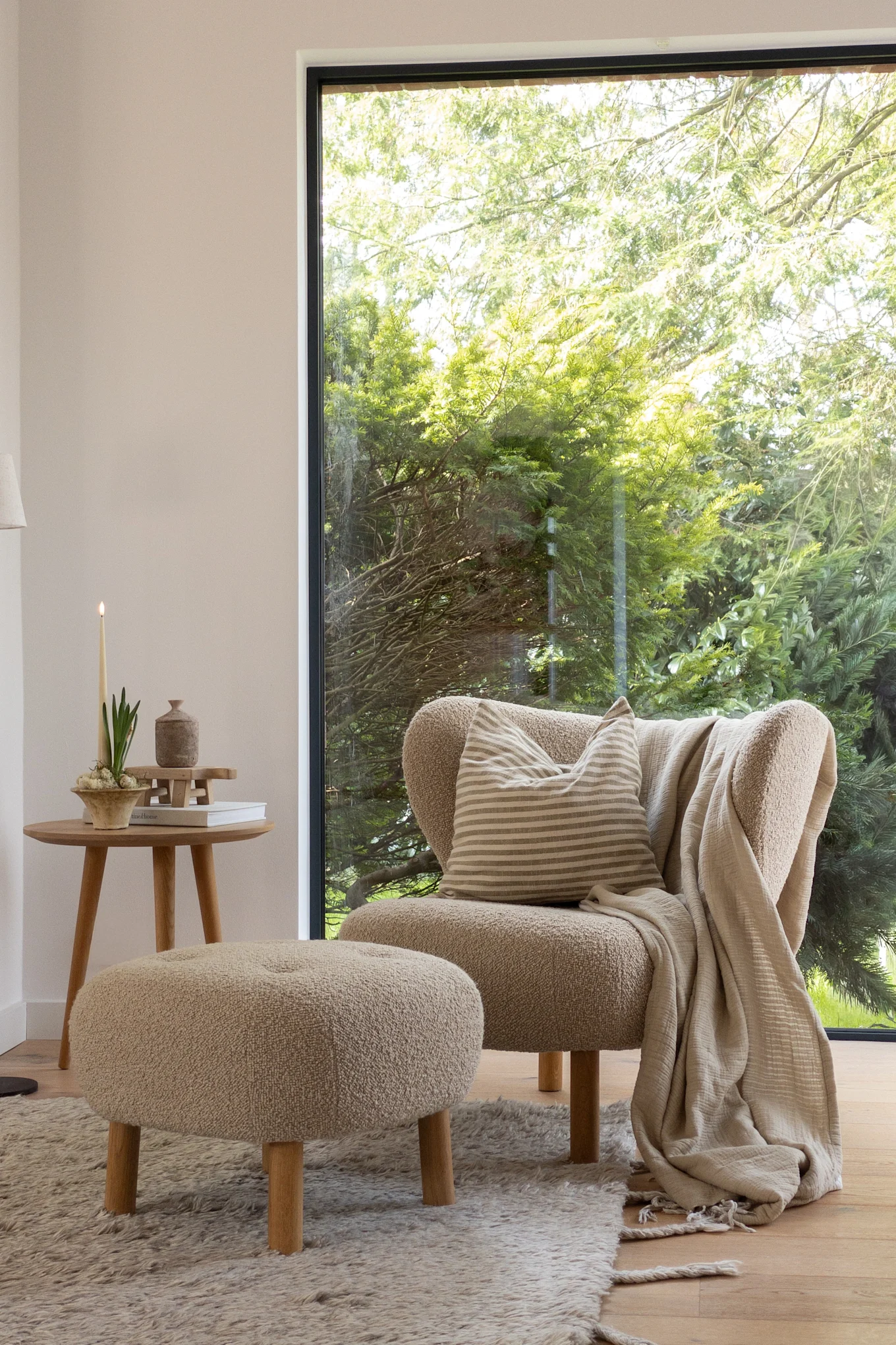 Gemütliches Wohnzimmer mit einem strukturierten beigen Sessel, passendem Hocker, rundem Beistelltisch aus Holz mit brennender Kerze und Pflanze sowie einem großen Fenster mit Blick auf einen grünen Garten.