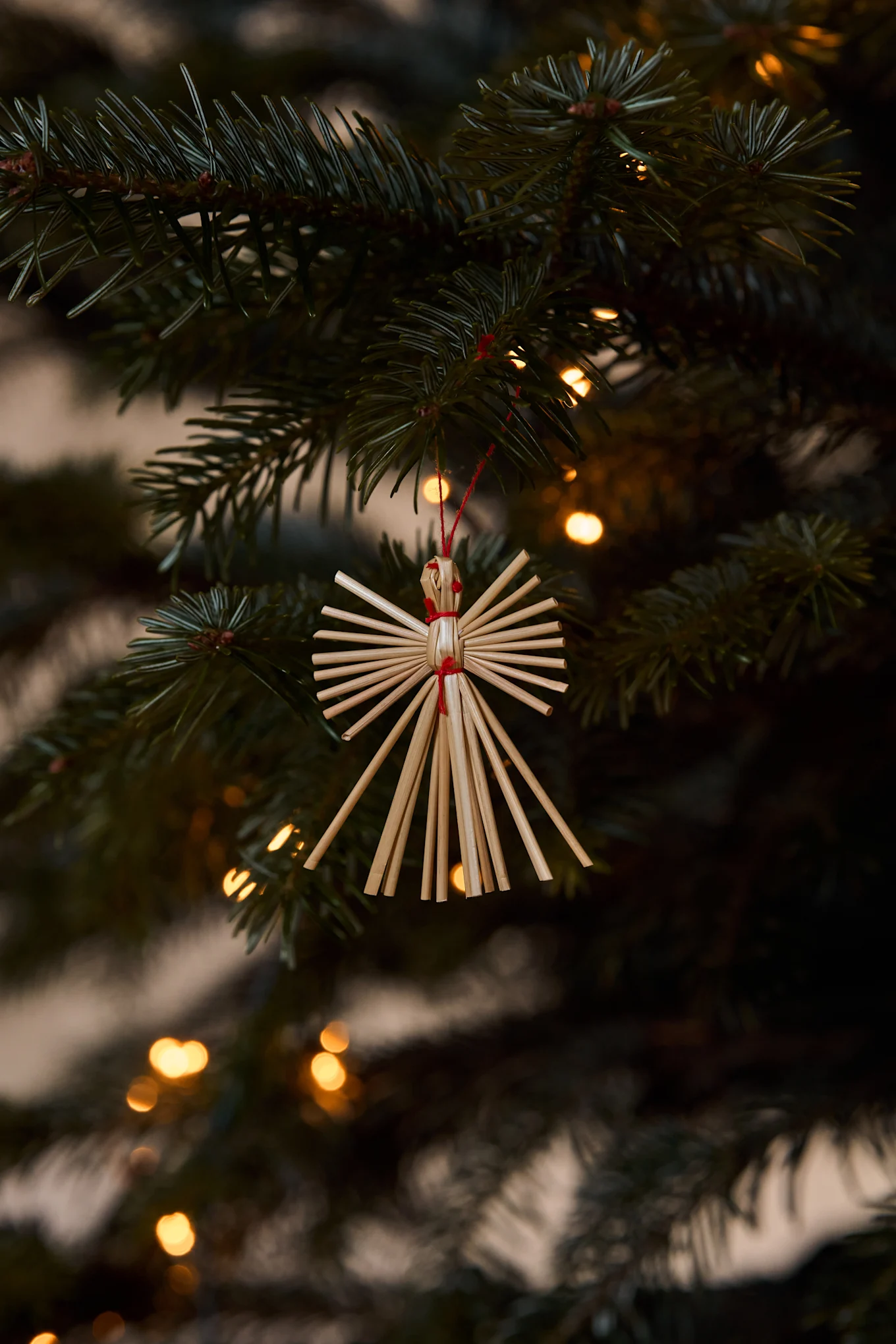 Strohengel-Ornament hängt an einem Weihnachtsbaumzweig mit verschwommenen Lichtern im Hintergrund.