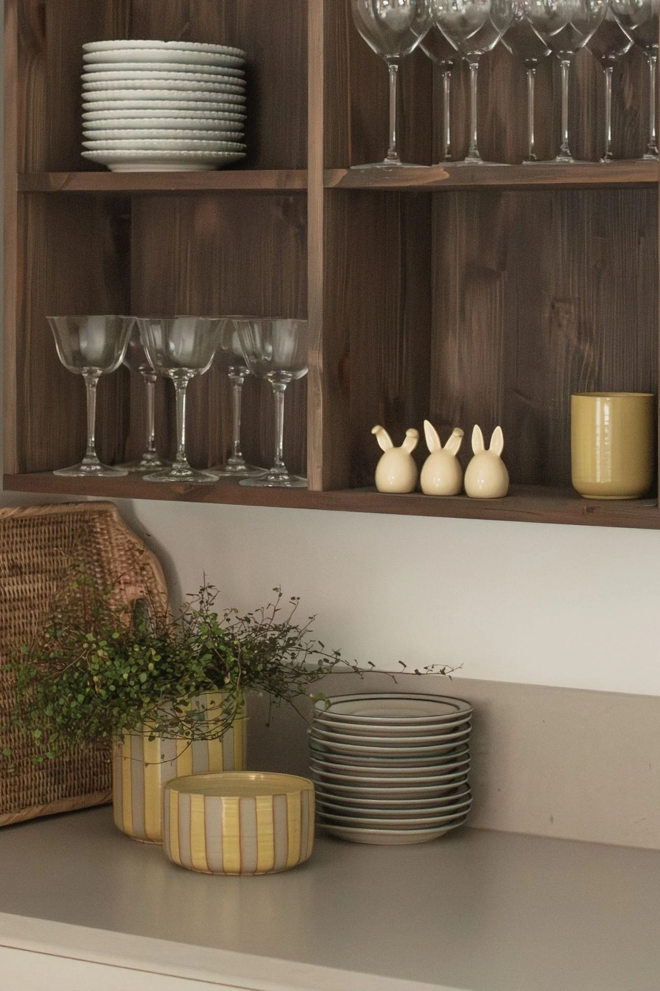 Holzregale mit gestapelten weißen Tellern, verschiedenen Gläsern, drei Keramikhasen und einem gelben Becher. Auf der Theke stehen ein geflochtener Korb, Pflanzen in gestreiften Töpfen und weitere Teller.