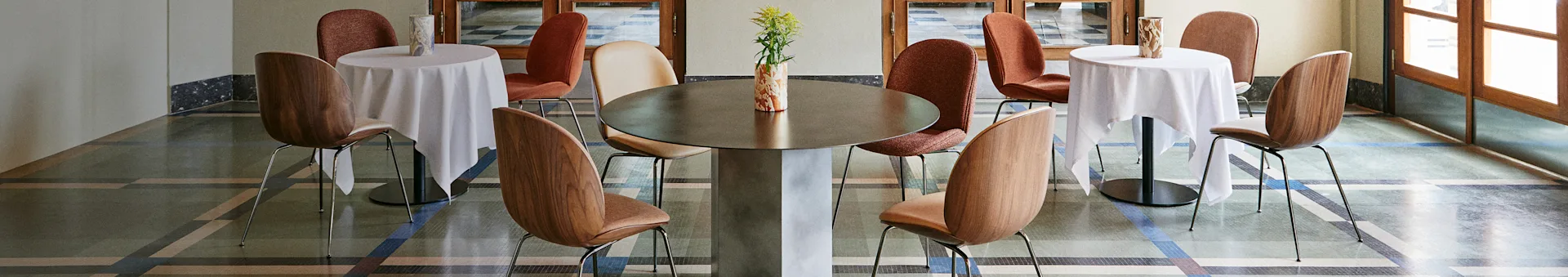 Stilvolles Café-Interieur mit einem großen zentralen Tisch, zwei kleineren Tischen mit weißen Decken und verschiedenen Stühlen auf einem gemusterten Boden.
