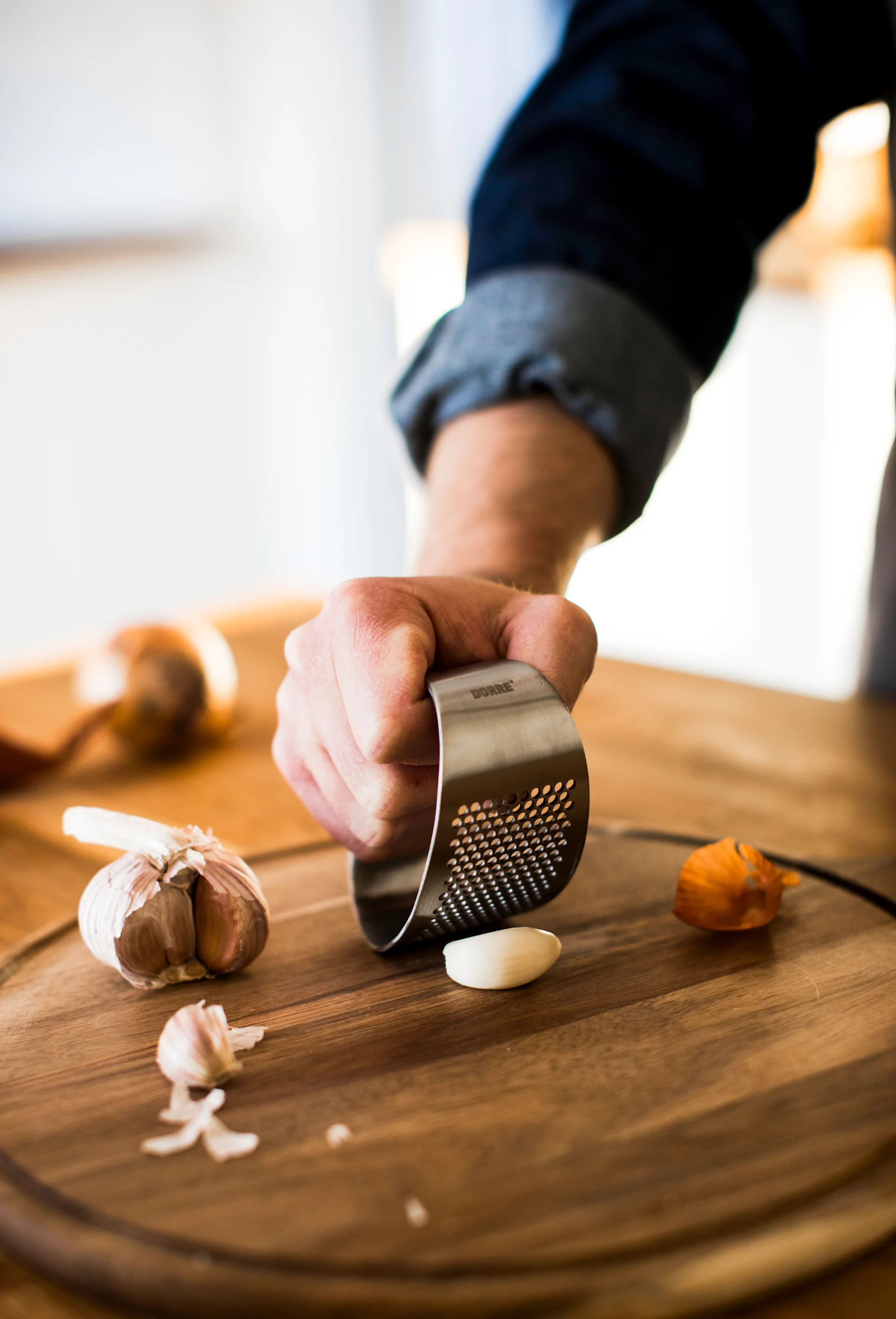Eine Hand presst eine Knoblauchzehe mit einer Edelstahl-Knoblauchpresse auf einem Holzschneidebrett.