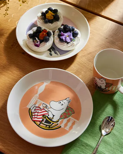 Moomintroll-Teller, Tasse und Löffel neben einem Teller mit Baisers mit Blaubeeren und Blumen auf einem Holztisch.