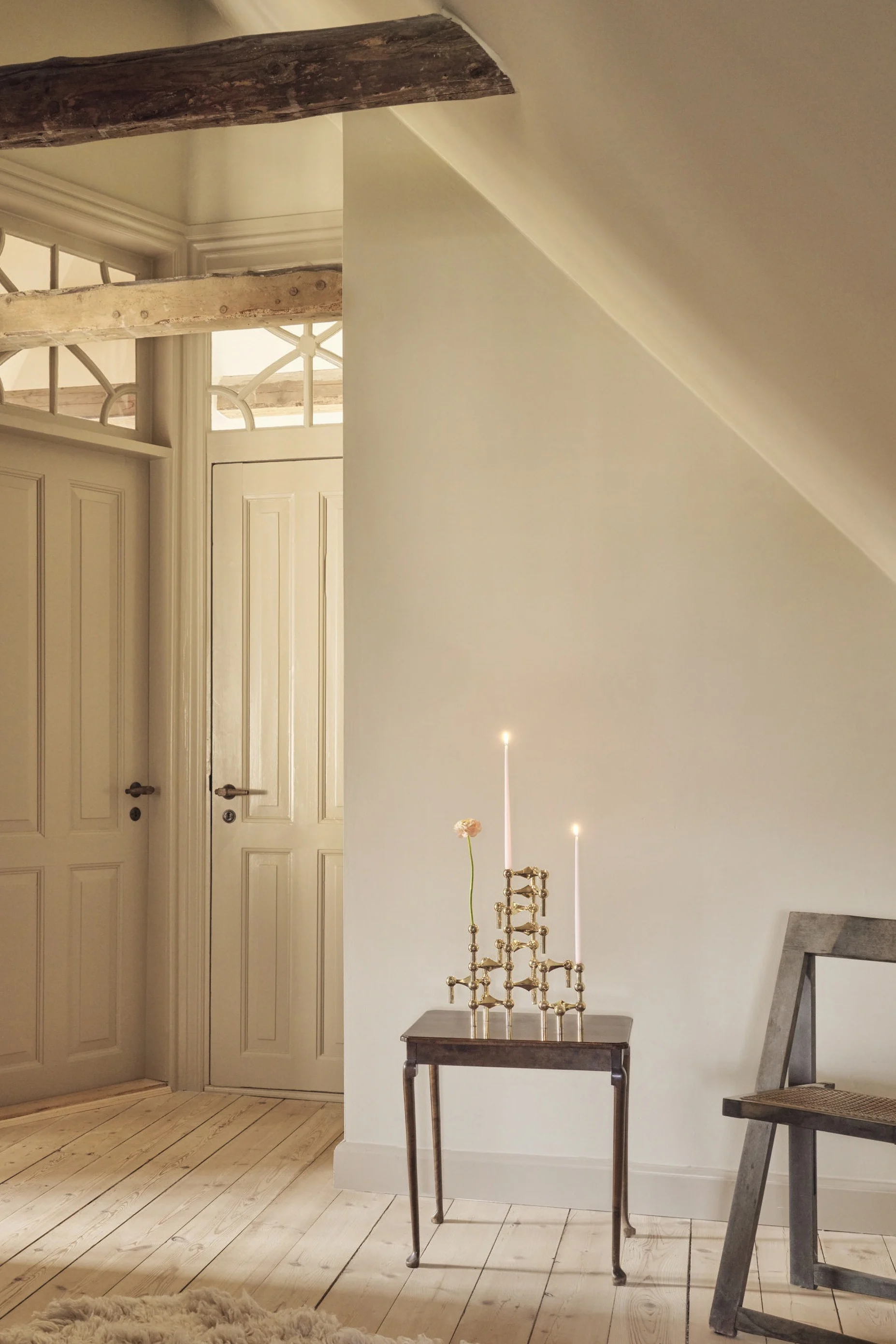 Ein Dachzimmer mit hellen Wänden und Holzböden. Zwei Kassettentüren mit verzierten Oberlichtern, ein sichtbarer Holzbalken, ein Beistelltisch mit brennenden Kerzen und einer Blume, ein Stuhl und ein flauschiger Teppich.