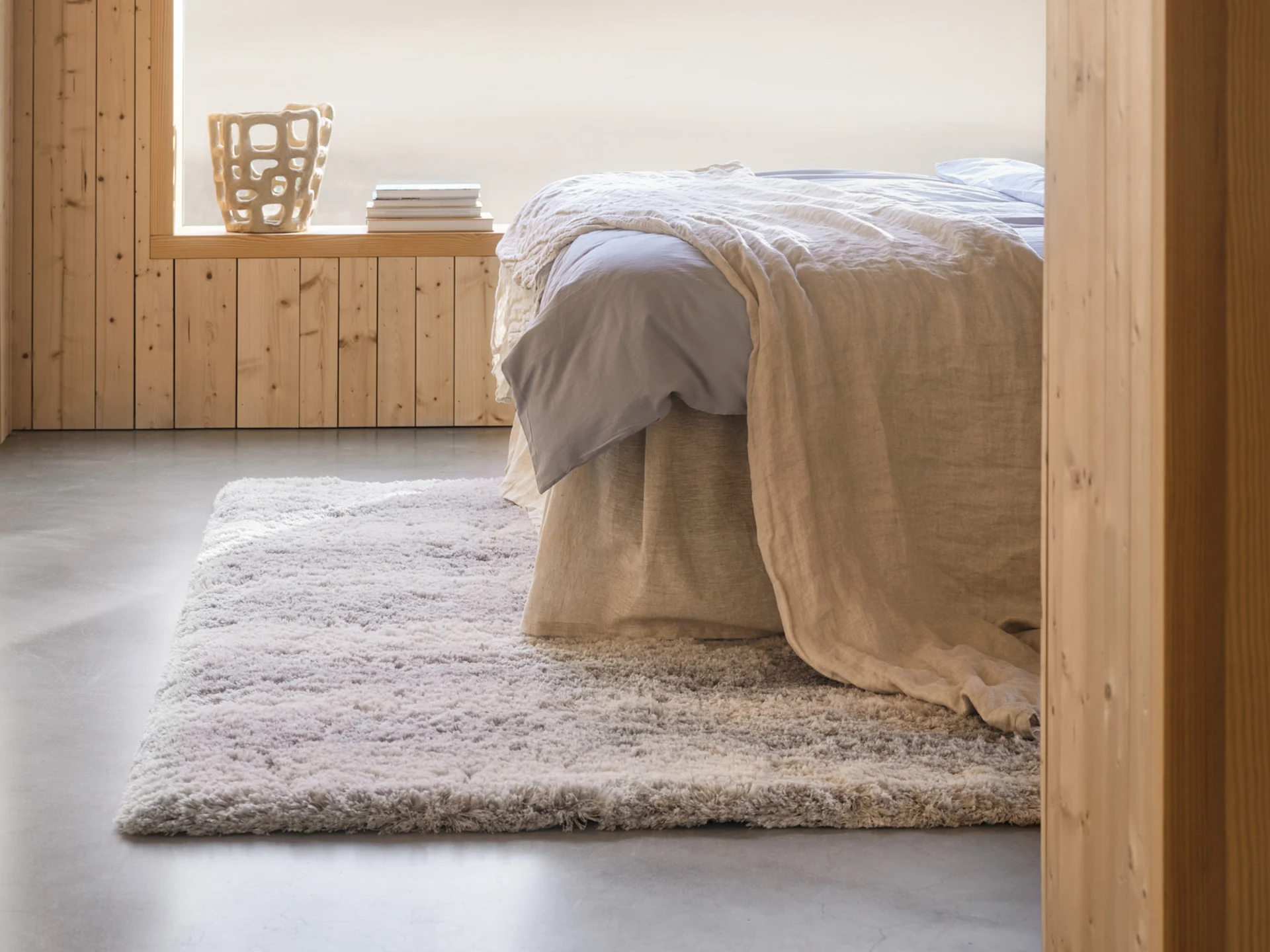 Gemütliches Schlafzimmer mit Bett, hellblauen Laken, beiger Decke, flauschigem Teppich, holzvertäfelten Wänden und einer Deko-Vase.