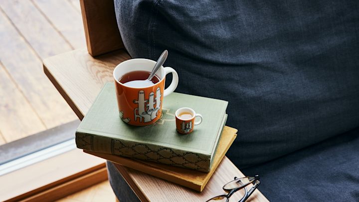 Die orangefarbene Mumin-Charaktertasse Hatifnatten mit Tee steht neben einem Sofa auf einem Stapel Büchern.