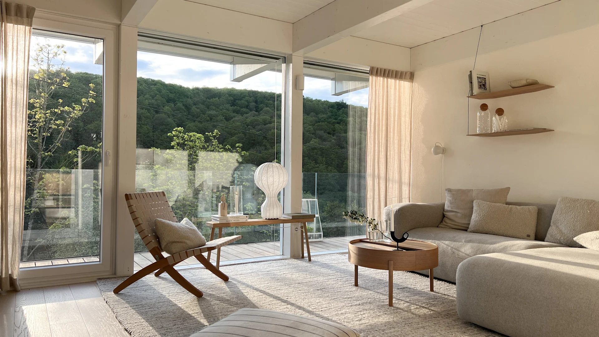 Ein helles Wohnzimmer mit großen Fenstern mit Blick auf grüne Hügel, mit einem beigen Ecksofa, einem runden Couchtisch aus Holz, einem gewebten Teppich, einem Leder- und Holzstuhl und einer skulpturalen weißen Lampe.