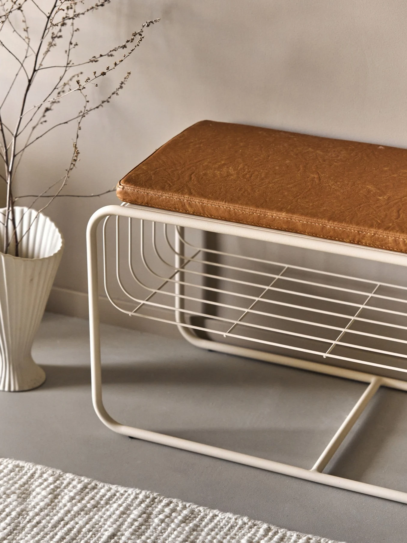 Moderne beige Metallbank mit braunem Kunstlederpolster und offener Lattenablage. Eine weiße Vase mit Ästen und ein Teppich sind in der Nähe.