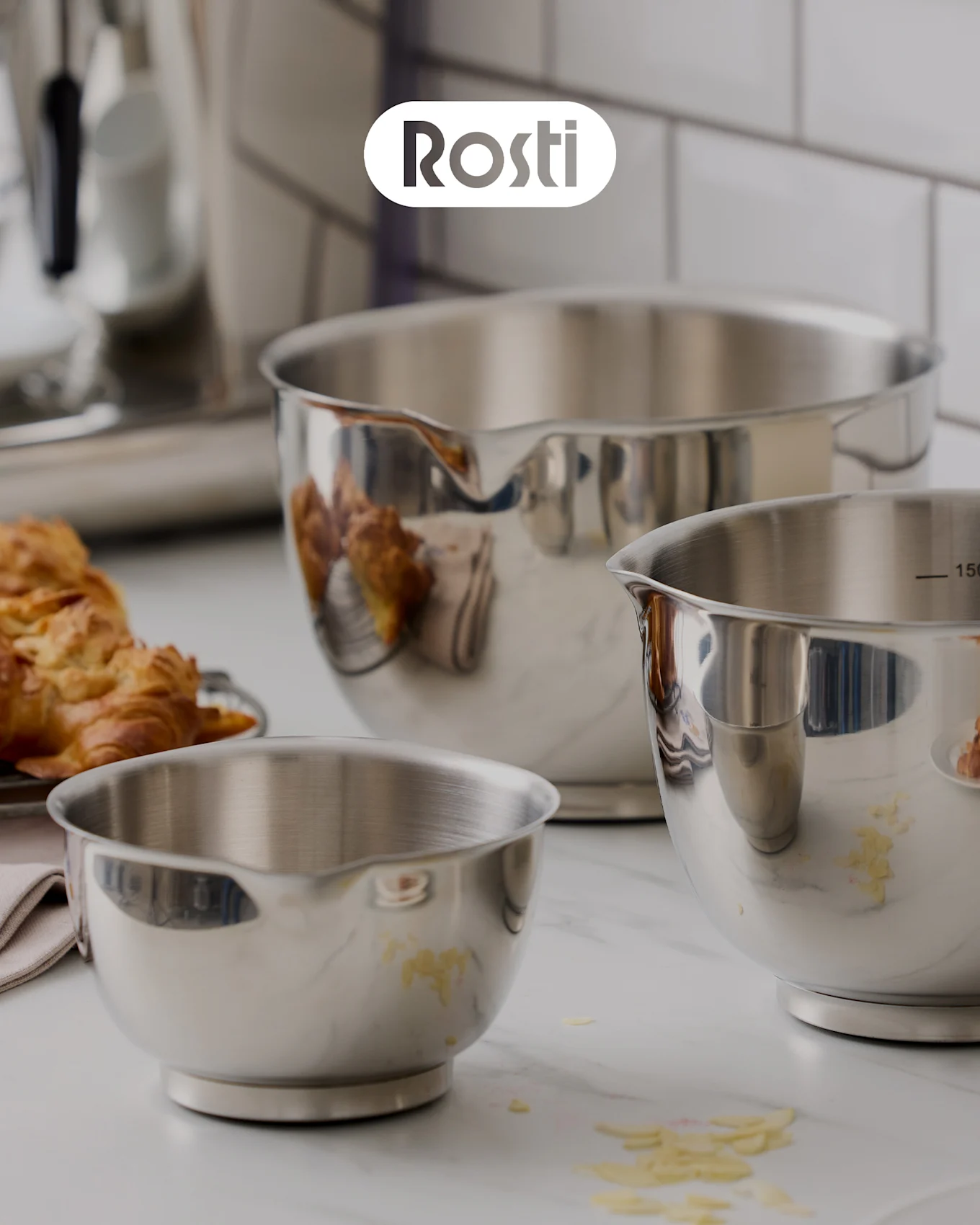 Drei Rosti-Schüsseln auf einer Theke: eine beige Schüssel mit einer blauen darüber, beide mit Deckeln, und eine kleine gelbe Schüssel gefüllt mit getrockneten Aprikosen.