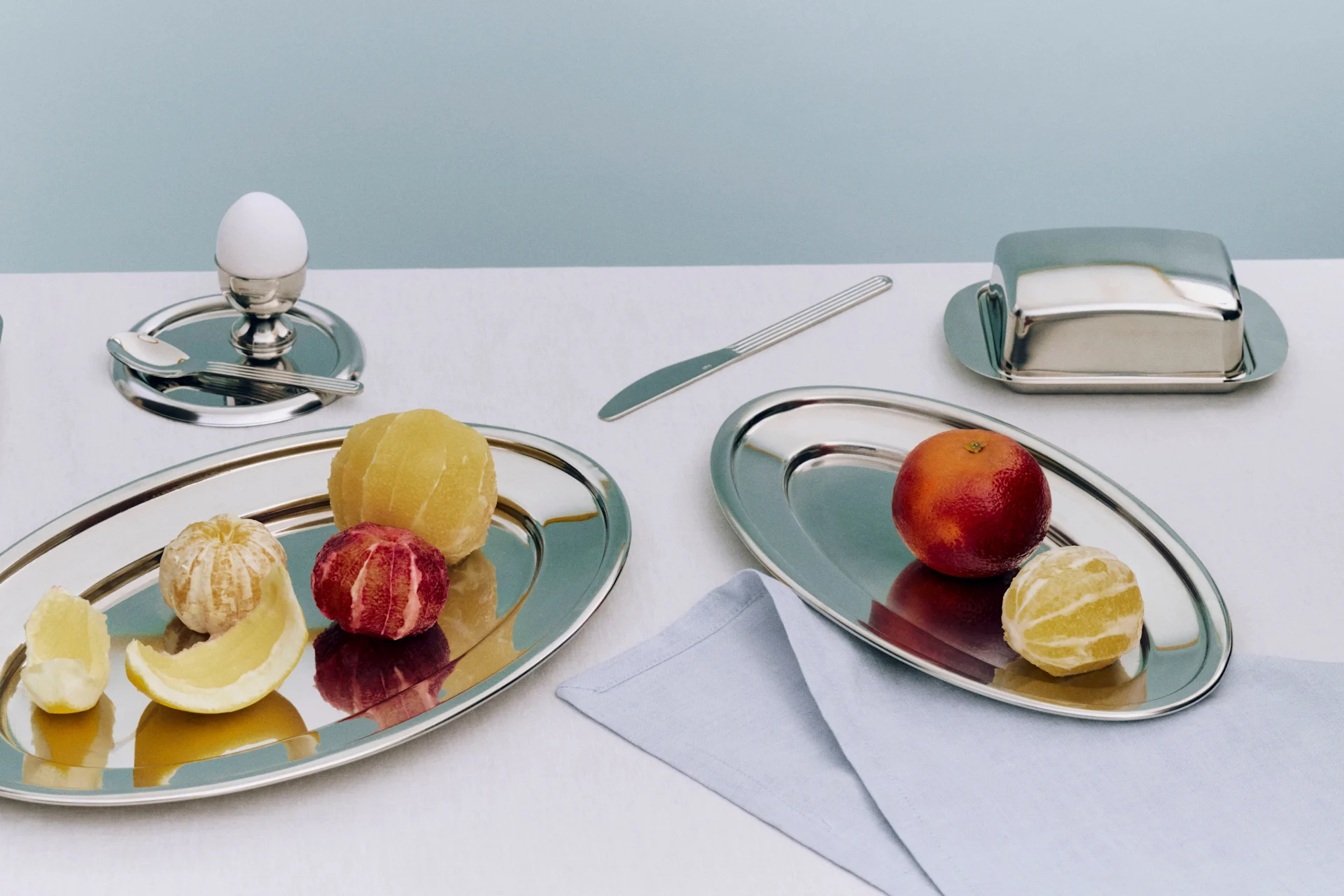 Ein Frühstücksgedeck mit zwei Silberschalen mit geschälten Zitrusfrüchten, einem Ei im Eierbecher, einer Butterdose und einem Messer.