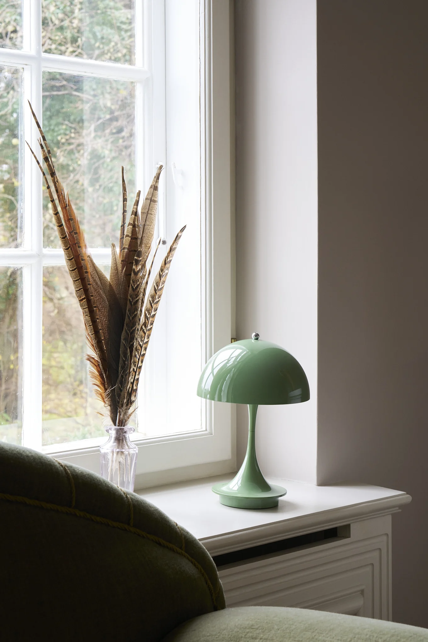 Eine grüne Pilzlampe und Feder-Vase auf einer Fensterbank neben einem grünen Sofa, mit einem Rattan-Krug im Hintergrund.
