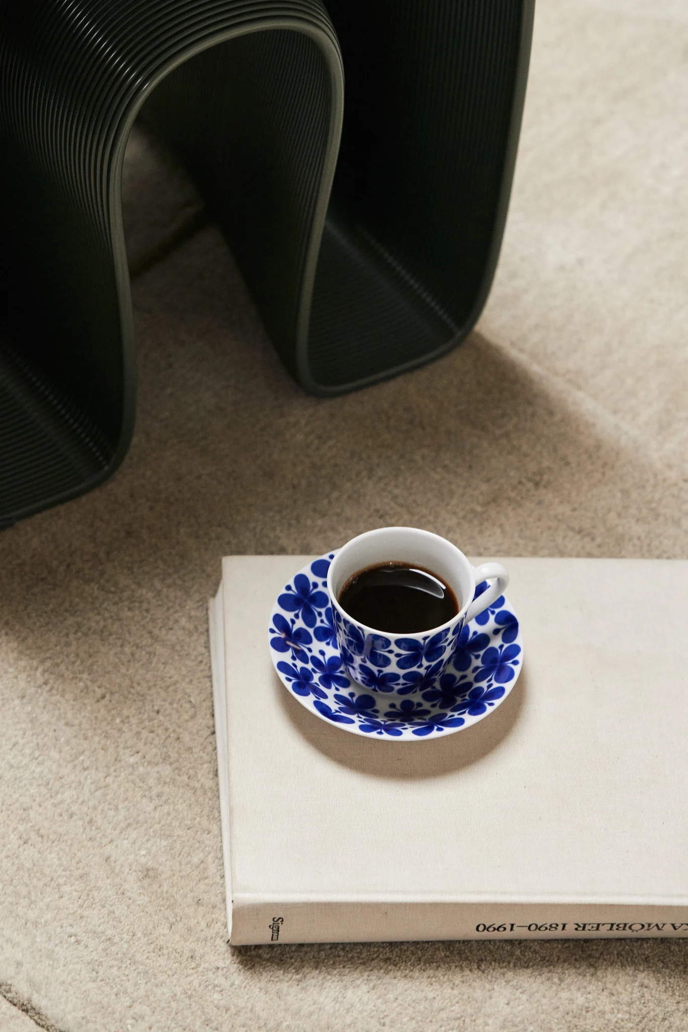 Kaffeetasse mit blauem Blumensaucer auf hellem Buch, dunkles geripptes Objekt im Hintergrund.