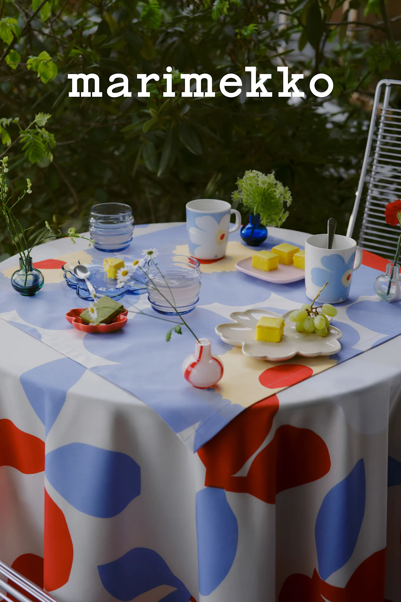 Marimekko-Logo. Ein gedeckter Tisch im Freien mit einer blau, rot und weiß gemusterten Tischdecke, Blumenbechern, gelben Kuchen, Trauben und kleinen Vasen.
