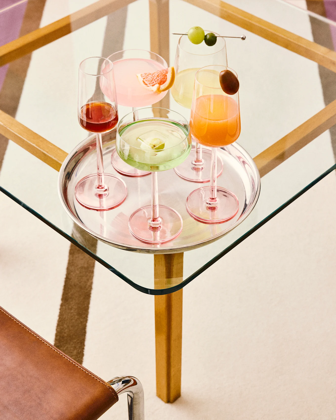 Fünf farbenfrohe Cocktails auf einem Silbertablett: pink mit Grapefruit, grün mit Fruchteis, gelb mit Trauben, orange mit Garnitur und dunkelrot, alle auf einem Glastisch.