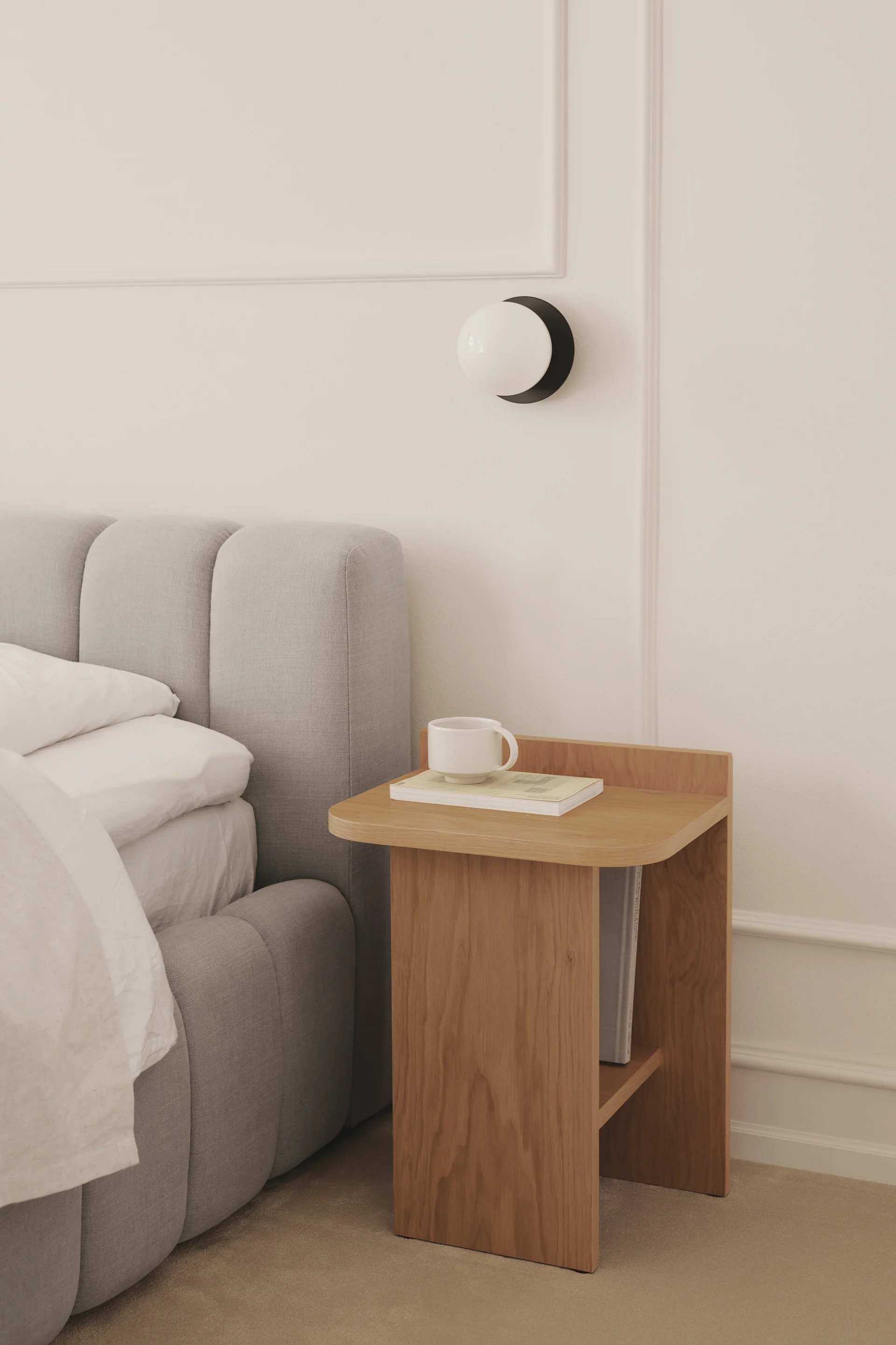 Ein gemütliches Schlafzimmer mit einem grauen Polsterbett neben einem Holznachttisch mit Tasse und Buch, sowie einer runden Wandlampe.