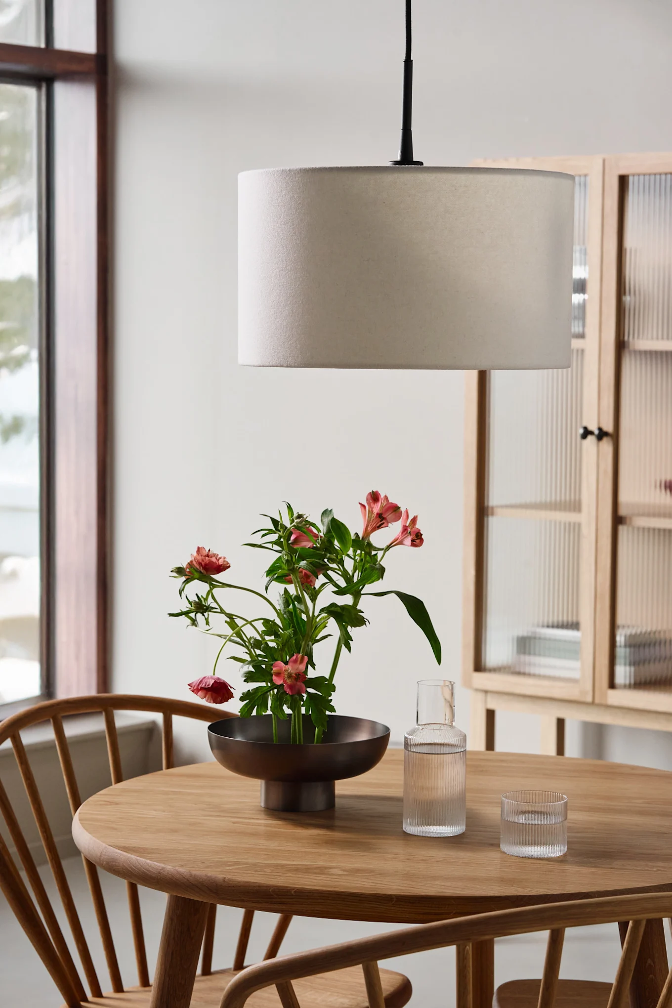 Ein runder Holztisch mit einer Schale rosa Blumen, einer Glaskaraffe, einem Glas und einer runden Pendelleuchte.