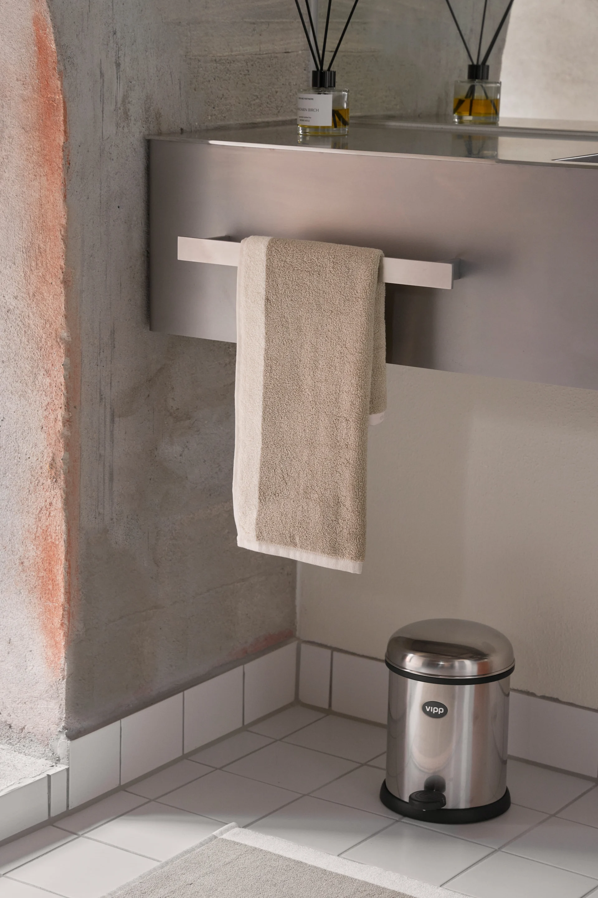 Modernes Badezimmer mit beigem Handtuch, einem Diffusor auf einer Metallablage und einem Treteimer aus Edelstahl.