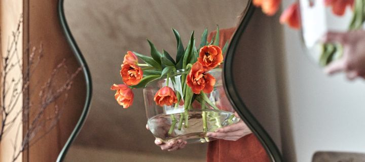 Frühlingserwachen: Ein Paar Hände trägt eine Vase mit frischen Blumen in einer klaren Glasvase, die sich in einem geschwungenen Spiegel von Ekbacken Studios spiegelt.