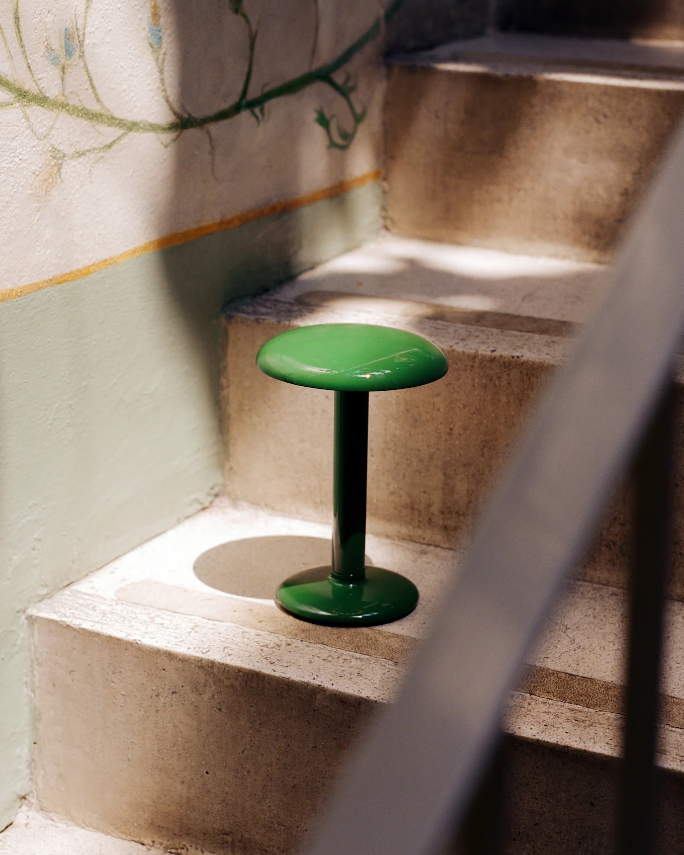 Ein glänzender, leuchtend grüner Hocker mit runder Platte und Sockel steht auf einer Betontreppe neben einer Wand mit einem verblassten Blumenwandbild.