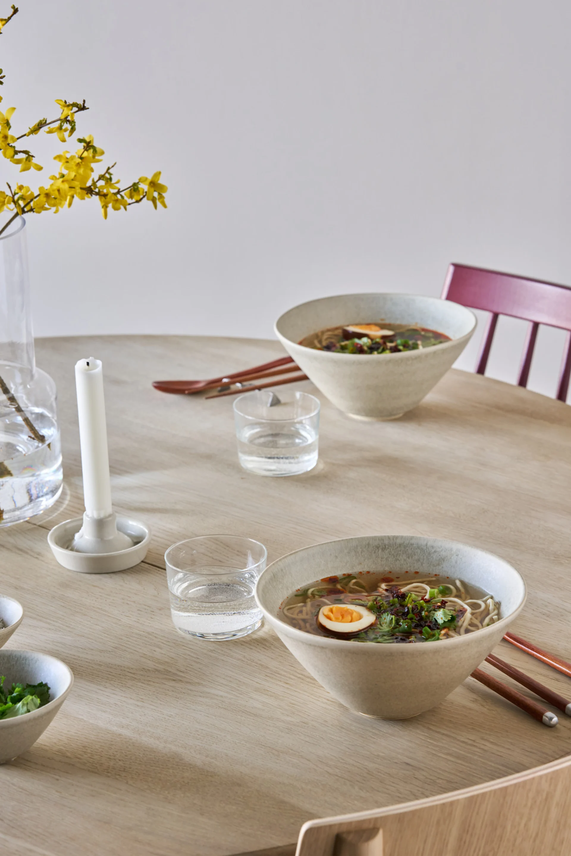 Zwei Schalen Ramen mit Eiern und Garnierung auf einem hellen Holztisch, mit Wassergläsern und gelben Blumen in einer Vase.