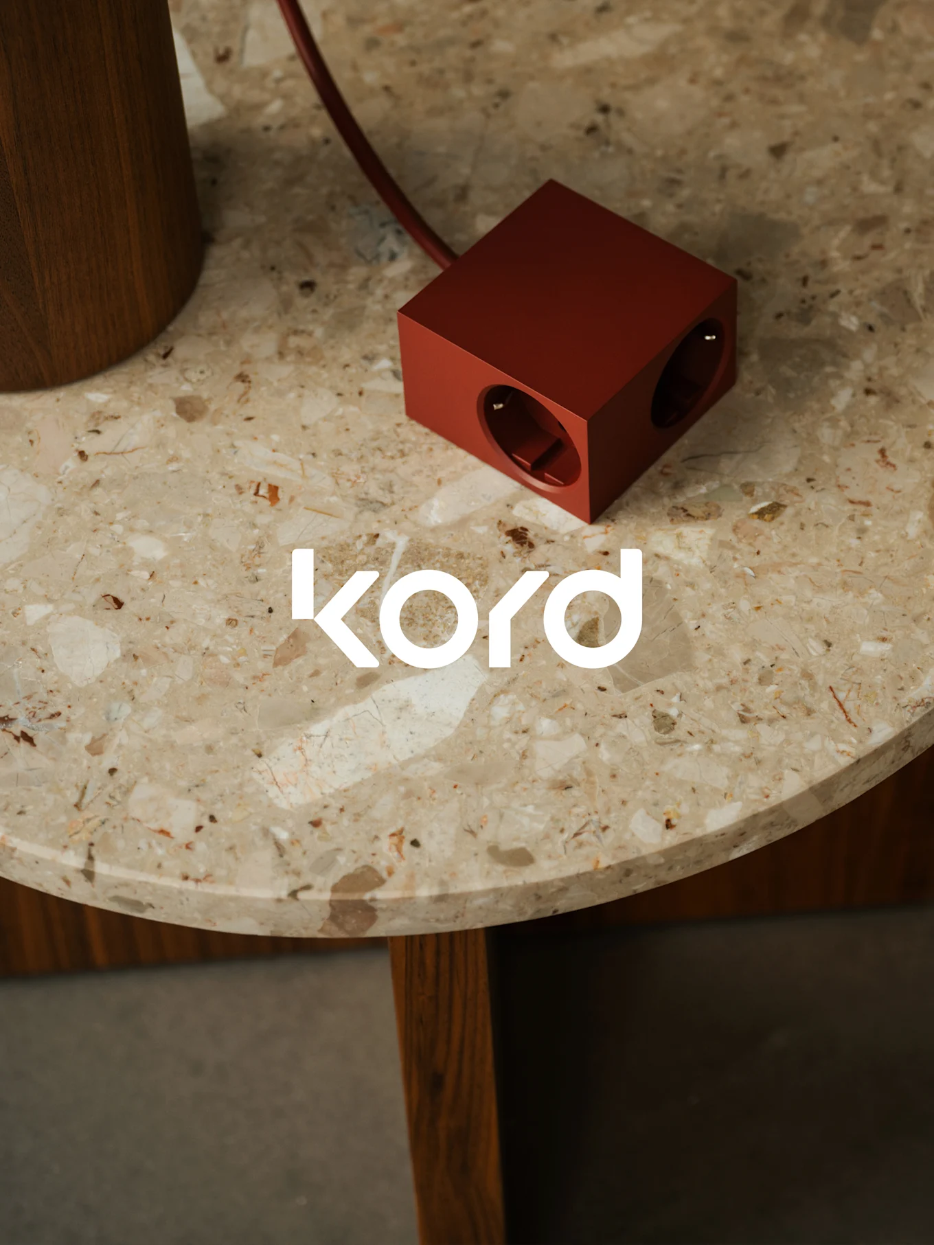Eine dunkelrote, würfelförmige Steckdosenleiste mit zwei sichtbaren Steckdosen liegt auf einem runden Marmortisch, mit dem weißen "kord"-Logo.