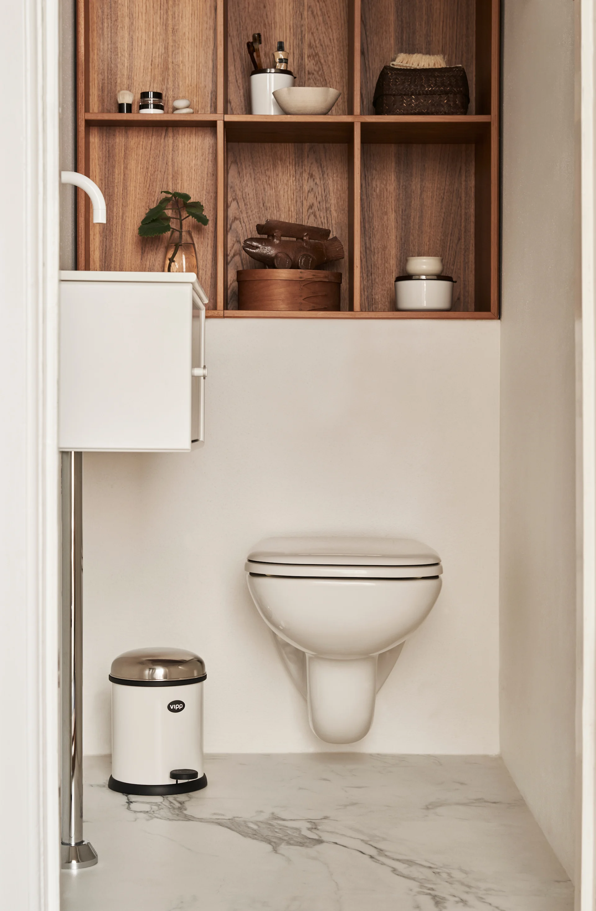 Modernes Badezimmer mit wandhängender Toilette, Waschbecken, einem weißen Vipp Treteimer und Holzregalen mit Toilettenartikeln.