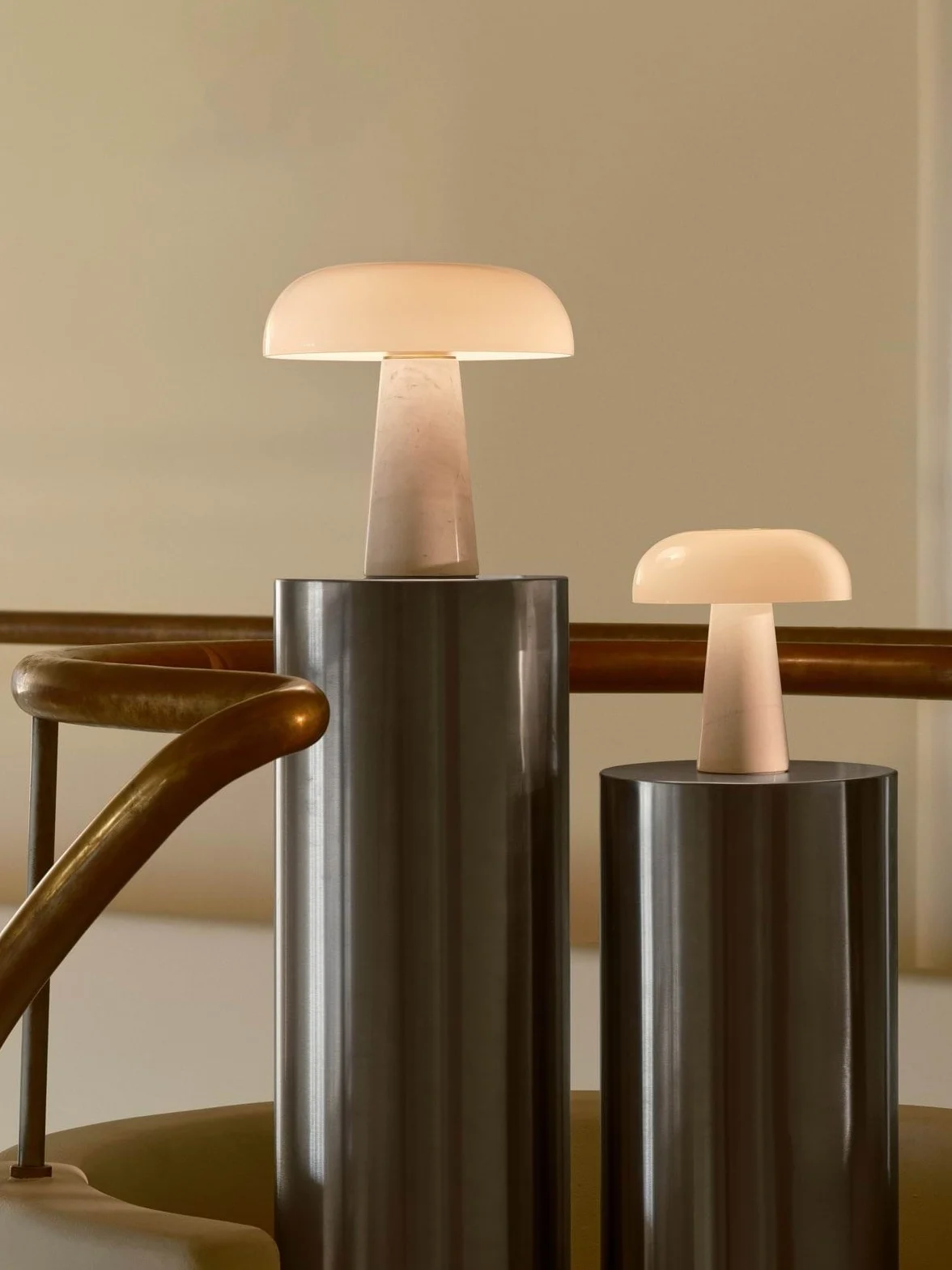 Zwei leuchtende pilzförmige Tischlampen, eine große und eine kleine, auf zylindrischen Podesten neben einem Messinggeländer.