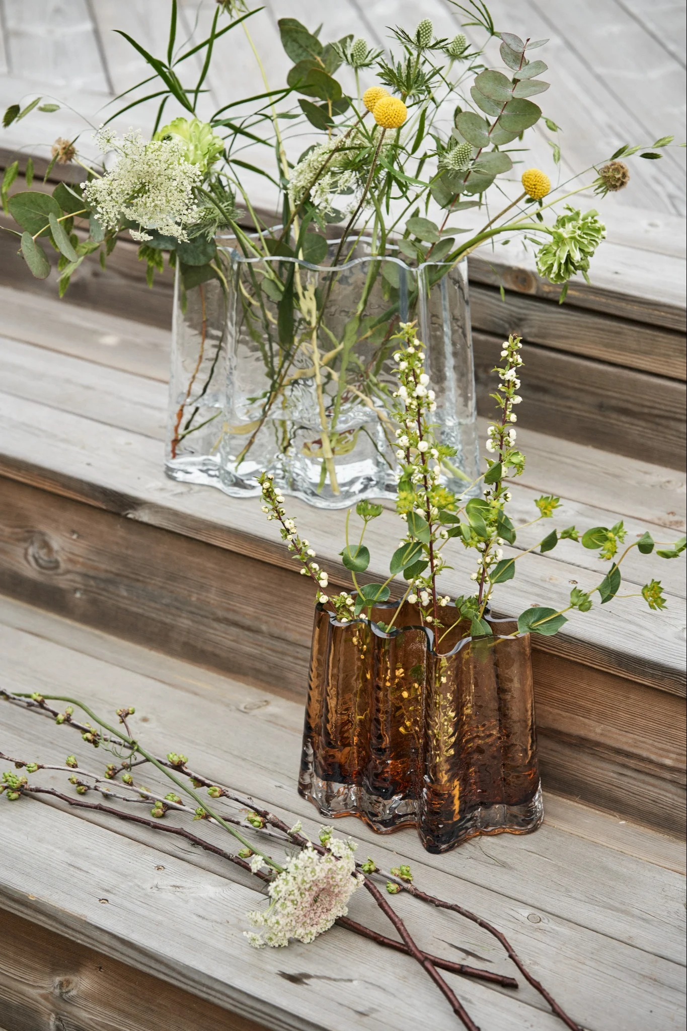 Eine durchsichtige und cognacfarbene Vase stehen auf einer Treppe aus Holz, umgeben von Frühlingsblumen. 