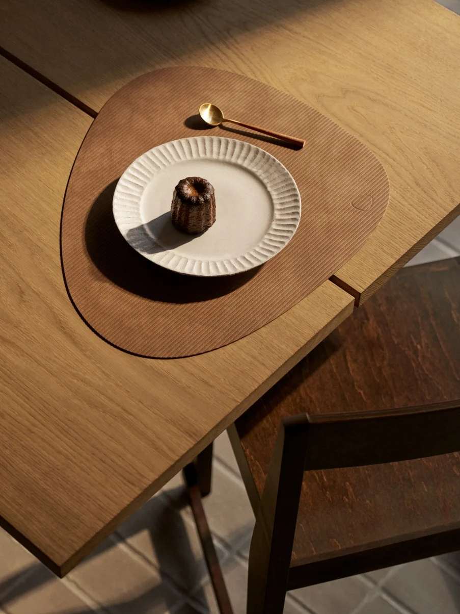 Aufnahme von oben: ein Canelé auf weißem Teller mit goldenem Löffel auf strukturierter brauner Unterlage auf Holztisch.