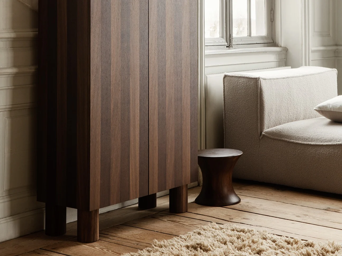 Eleganter Raum mit geriffeltem Schrank aus dunklem Holz, kniender Frauenskulptur, hellem Modulsofa und großem Fenster.