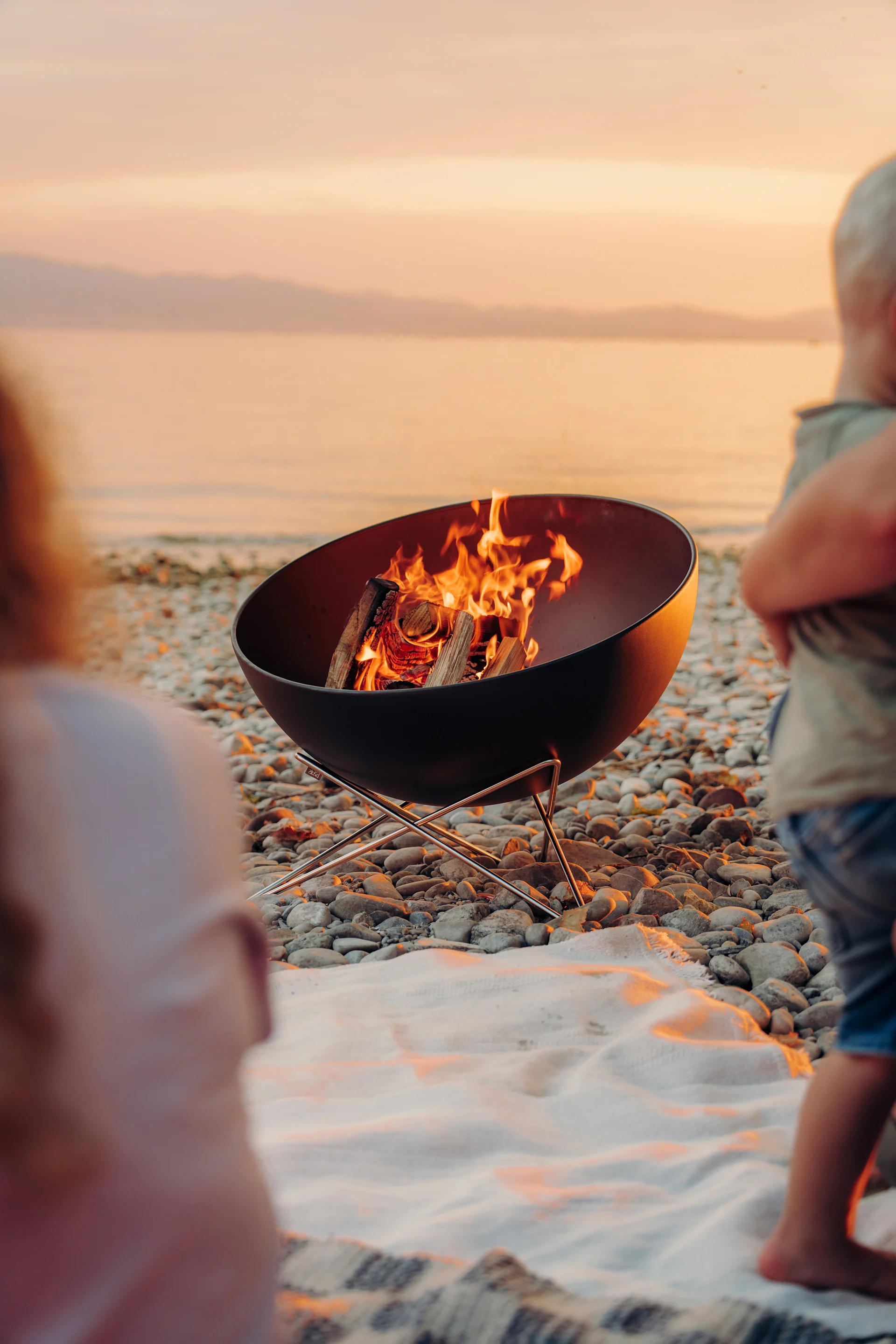 Eine Feuerschale mit glühenden Flammen an einem Kiesstrand bei Sonnenuntergang, mit unscharfen Personen im Vordergrund.