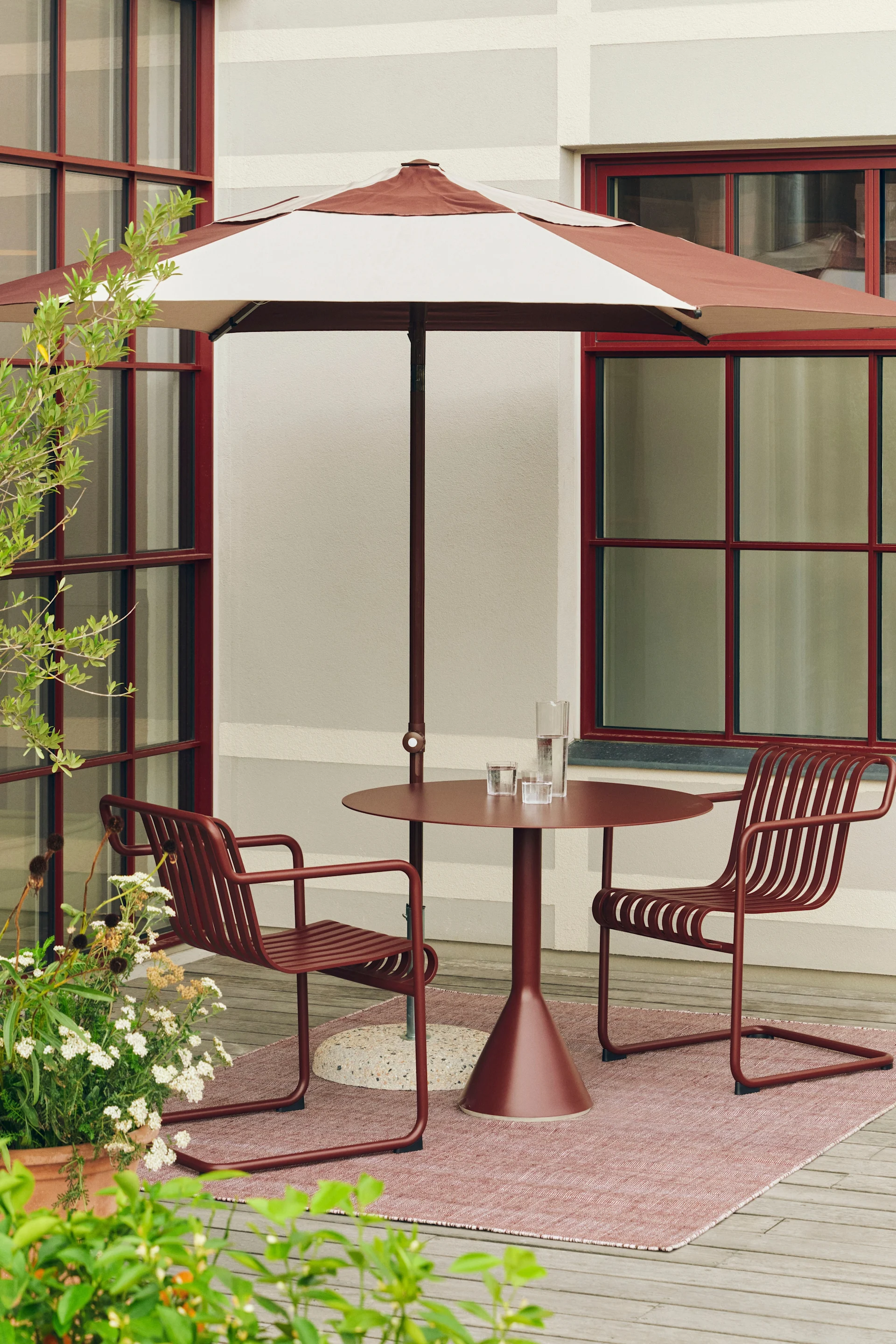 Gartenterrasse mit rot-braun gestreiftem Sonnenschirm, passendem Rundtisch und zwei Stühlen auf rosa Teppich.