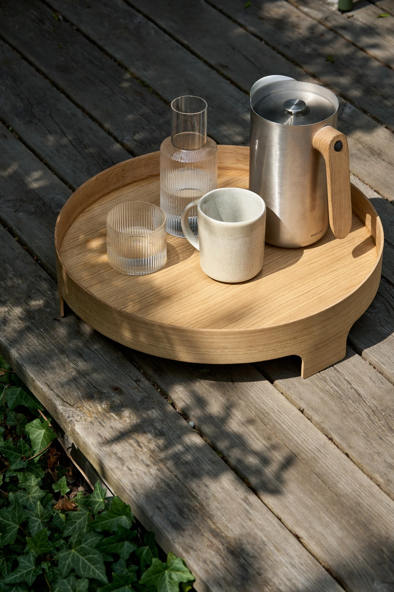 Das Bridge Tablett von Design House Stockholm steht draußen auf einer Terrasse, darauf die Nordic Kitchen Kanne von Eva Solo, ein Glas und die Karaffe aus der Ripple-Kollektion von Ferm Living, sowie die Sandsbro-Tasse von Scandi Living.