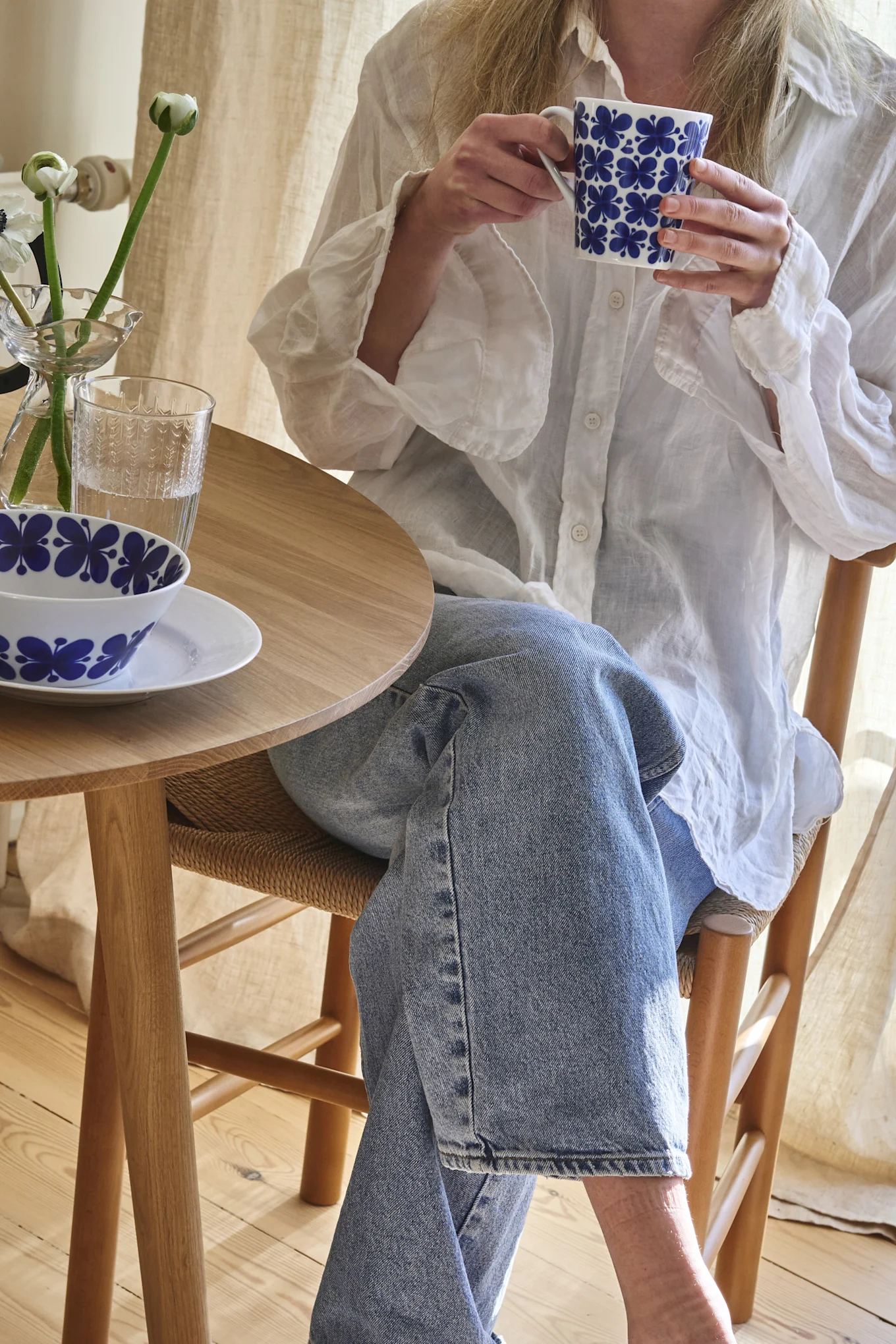 Person in weißem Hemd und Jeans hält eine blau geblümte Tasse an einem Holztisch mit passender Schale und Blumen.