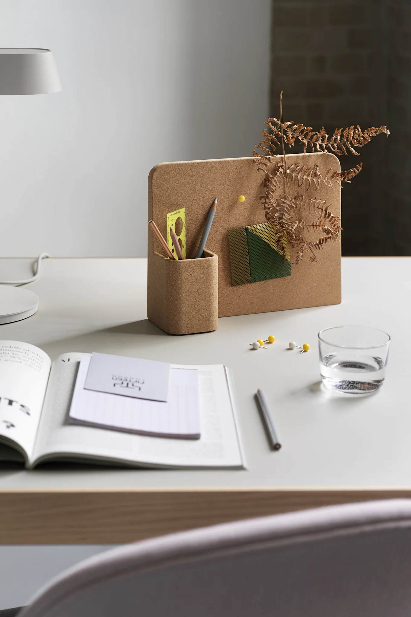 Kork-Memoboard mit integriertem Stiftehalter, gefüllt mit Bleistiften, einem Lineal und einem dekorativen Farn. Ein offenes Buch, ein Wasserglas und Reißnägel liegen auf dem weißen Schreibtisch.