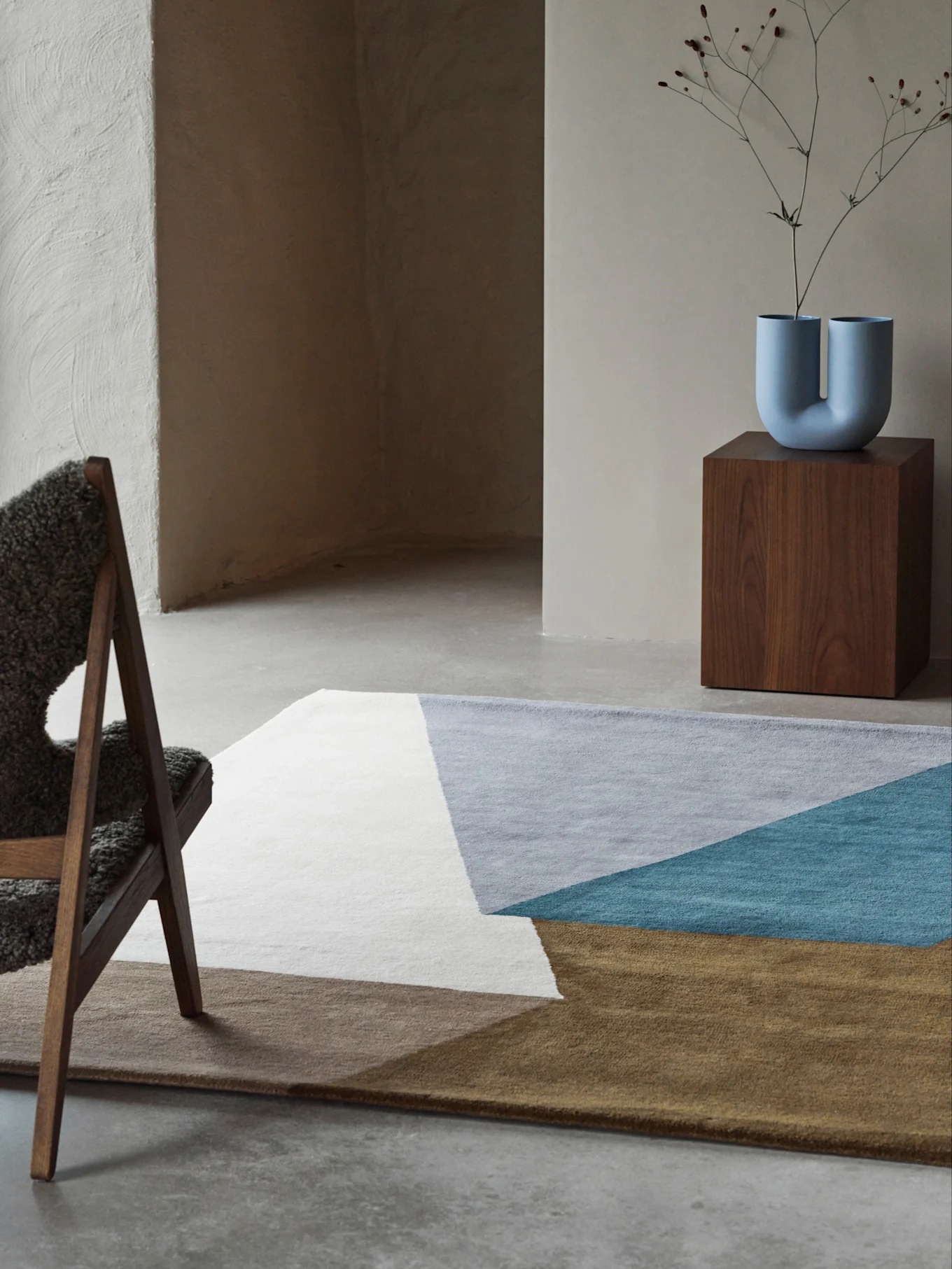 Modernes Zimmer mit einem geometrischen Teppich in Weiß, Grau, Türkis und Braun, einer U-förmigen blauen Vase auf einem Holzsockel und einem flauschigen Stuhl.