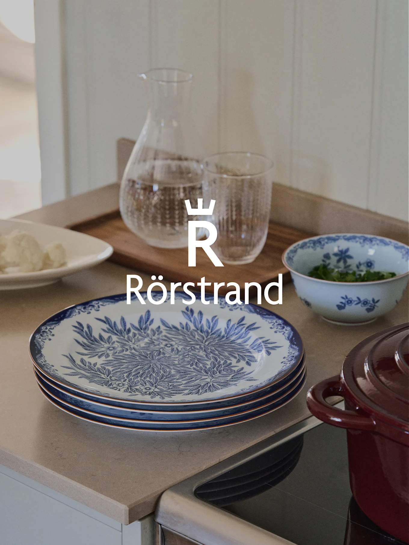 Rörstrand-Logo über Küchentheke mit blau-weißen Tellern, Karaffe, Glas und rotem Topf.
