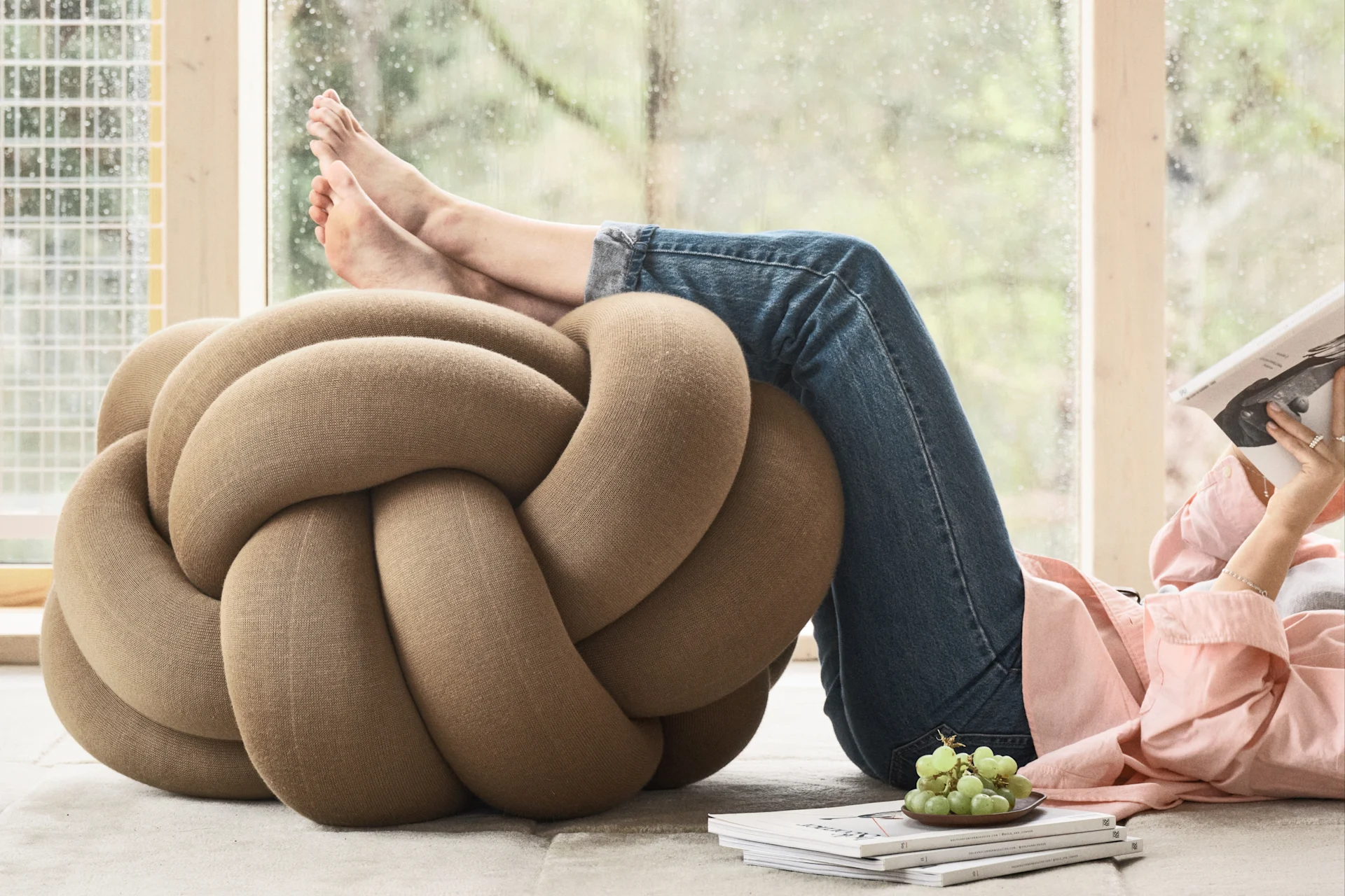 Die Knotenkissen von Design House Stockholm bietet auch den optimalen Komfort. Hier sehen Sie das beigefarbene Knot-Kissen in XL auf dem Fußboden liegen, eine auf dem Boden liegende Person hat ihre Beine darauf platziert.
