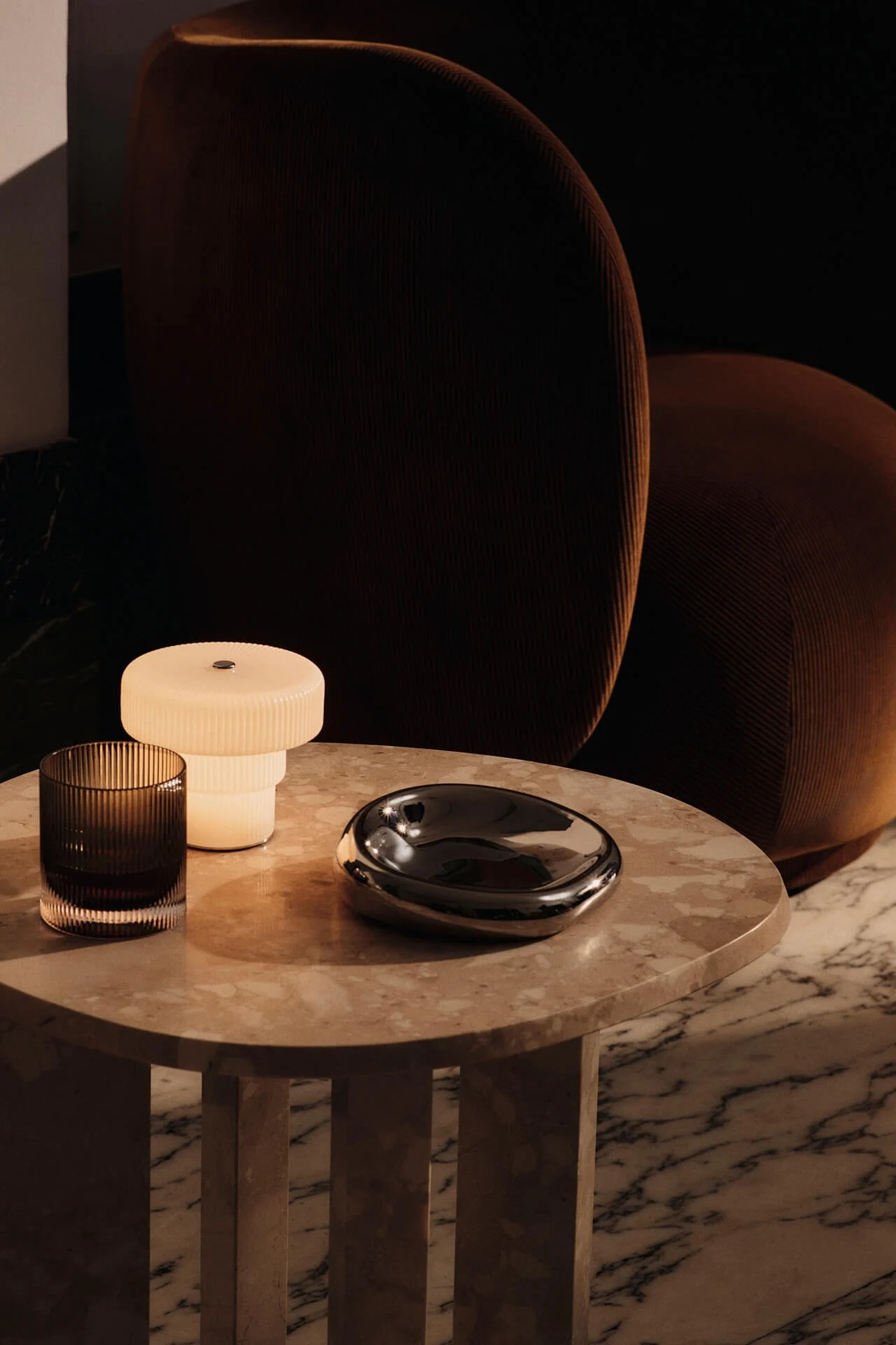 Die Akku-Tischleuchte Ripple von Ferm Living steht auf einem Beistelltisch aus Stein, neben dem ein Rico-Sessel steht.