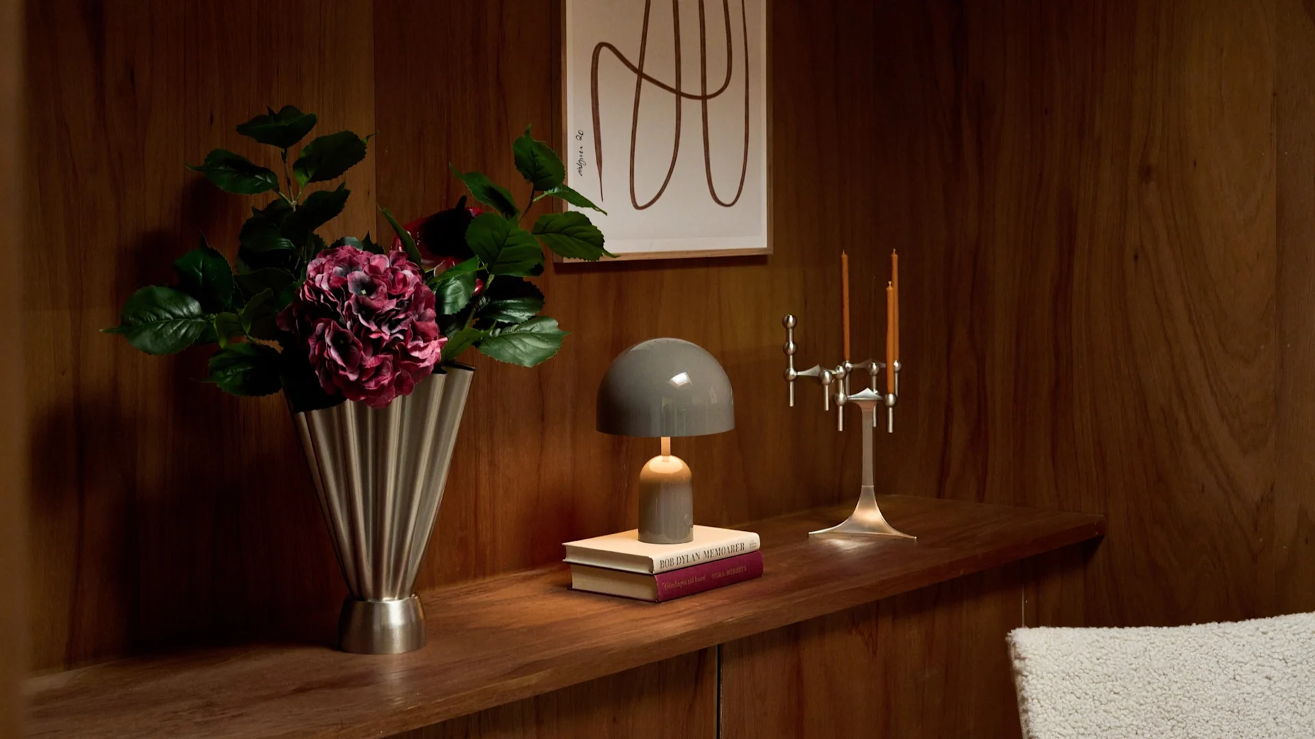Vase mit dunkelrosafarbenen Blumen, graue Pilzlampe auf Büchern, silberner Kerzenleuchter auf Holzregal mit Kunst.