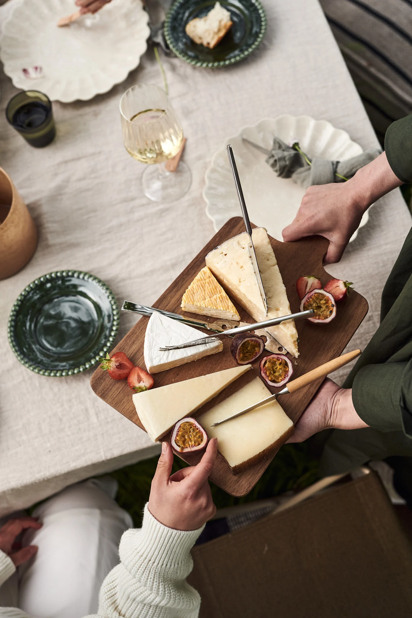 Hände servieren ein Holzkäsebrett mit verschiedenen Käsesorten, halbierten Erdbeeren und Passionsfrüchten auf einem gedeckten Tisch.