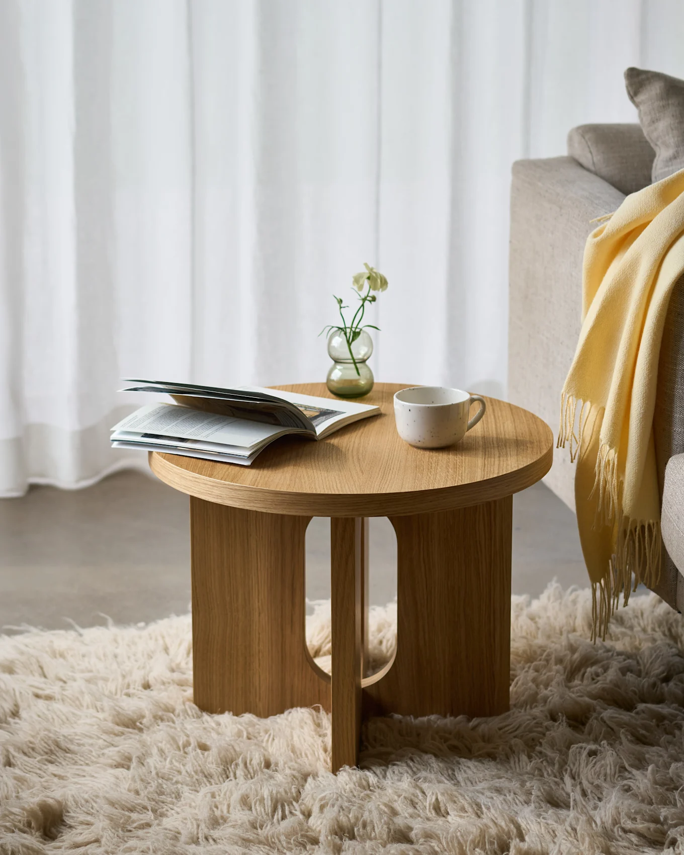 Runder Holznebentisch auf flauschigem Teppich, darauf ein offenes Magazin, eine weiße Blume in einer Vase und eine Keramiktasse, neben einem Sofa.
