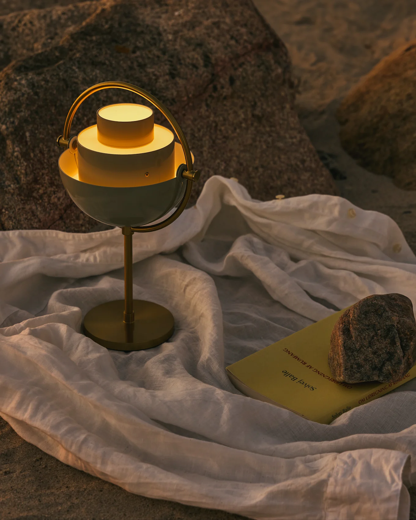 Eine moderne weiße und messingfarbene Tischlampe mit warmem Licht, einem gelben Buch und einem Stein auf einem weißen Tuch auf felsigem Grund.