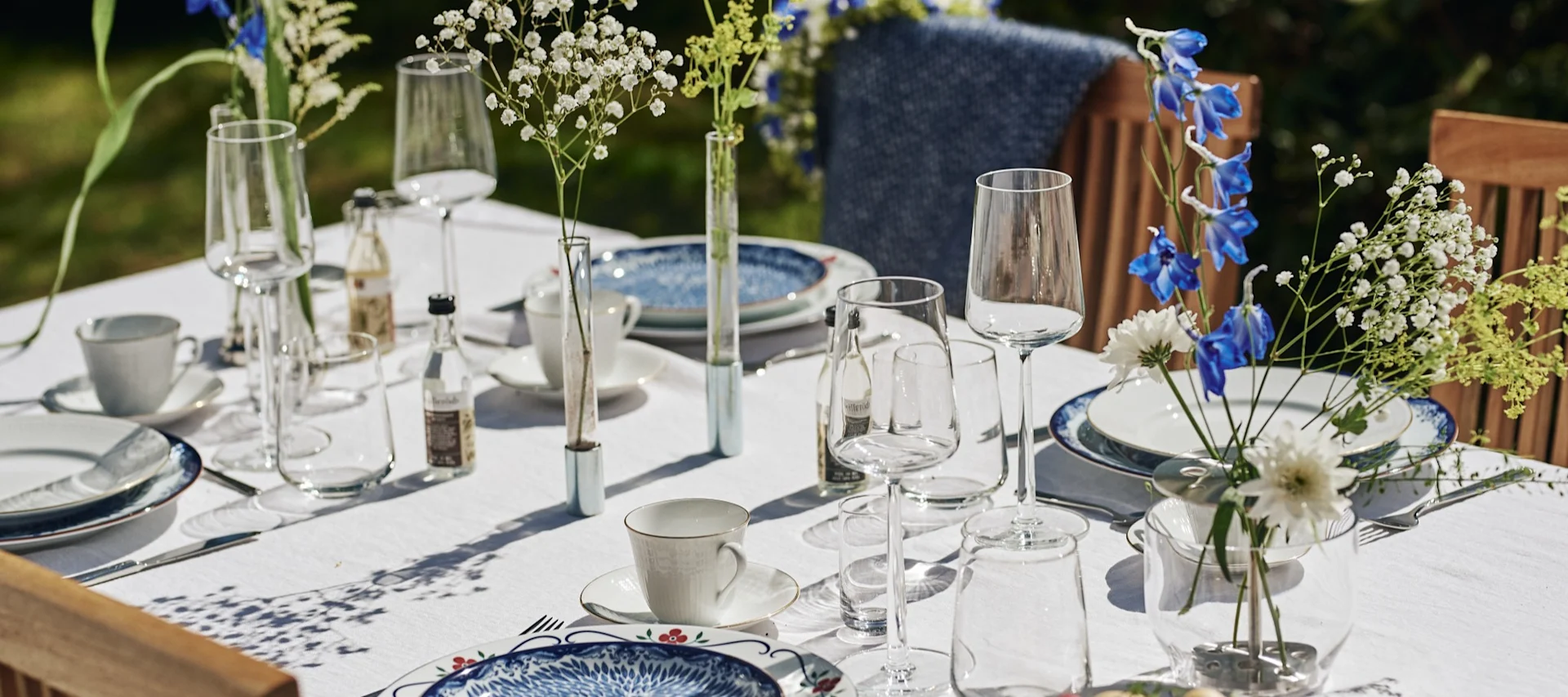 Midsommar Tischdeko: Die klassische Tischdeko in Schweden ist blau-weiß. Hier sehen Sie einen großen Tisch im Garten mit weißer Tischdecke, blau-weißem Geschirr und vielen kleinen Vasen mit Wiesenblumen.
