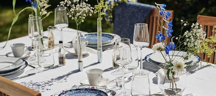 Midsommar Tischdeko: Die klassische Tischdeko in Schweden ist blau-weiß. Hier sehen Sie einen großen Tisch im Garten mit weißer Tischdecke, blau-weißem Geschirr und vielen kleinen Vasen mit Wiesenblumen.