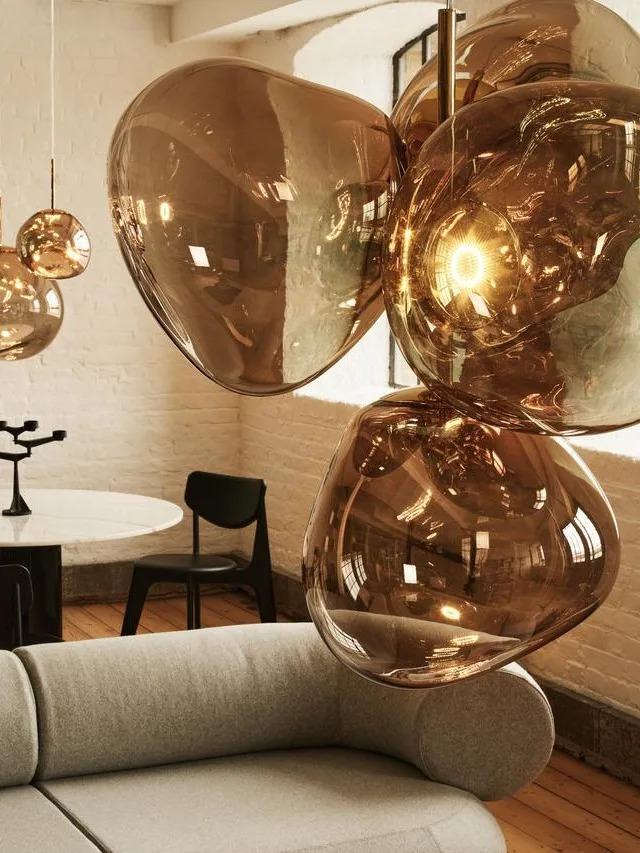 Ein modernes Wohnzimmer mit einem geschwungenen grauen Sofa, einem passenden Ottoman mit Zeitschriften und bernsteinfarbenen Glaspendelleuchten.