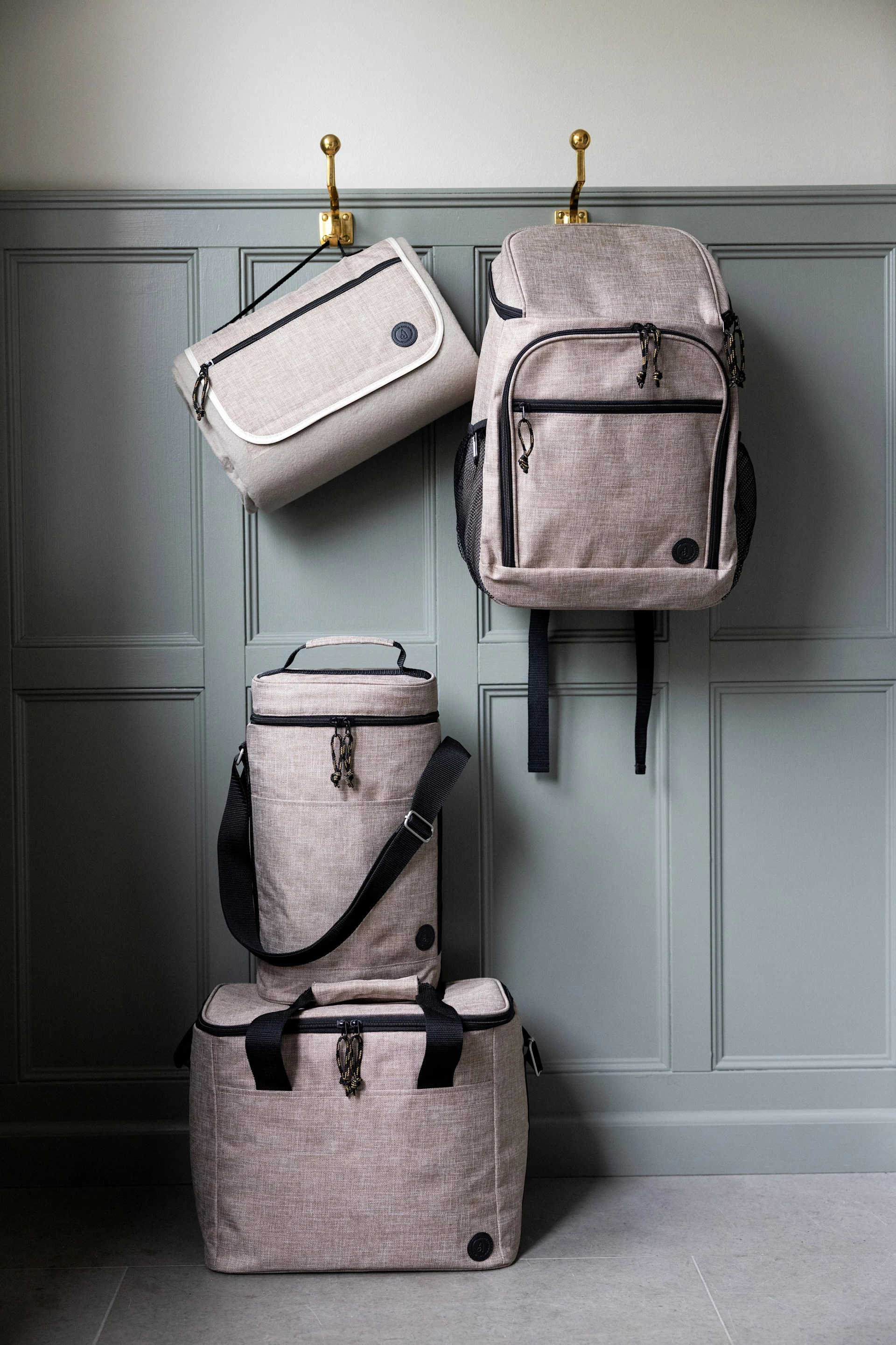 Vier hellbeige Taschen: eine flache Tasche und ein Rucksack hängen an Haken, eine zylindrische und rechteckige Kühltasche stapeln sich darunter.