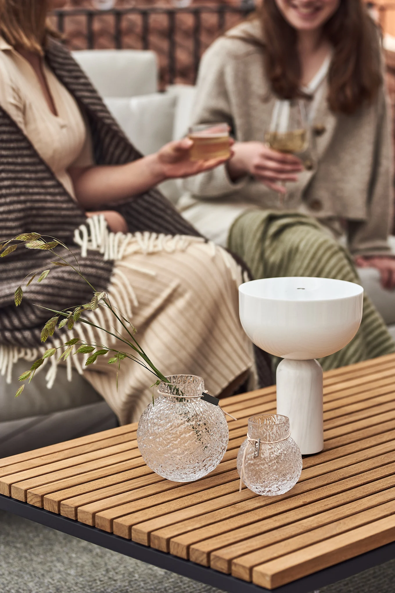Zwei Frauen sitzen auf einem Sofa auf dem Balkon und stoßen an. Im Vordergrund stehen die kabellose Kizu Tischleuchte von New Works und Vasen von Ernst in verschiedenen Größen auf einem Holztisch.